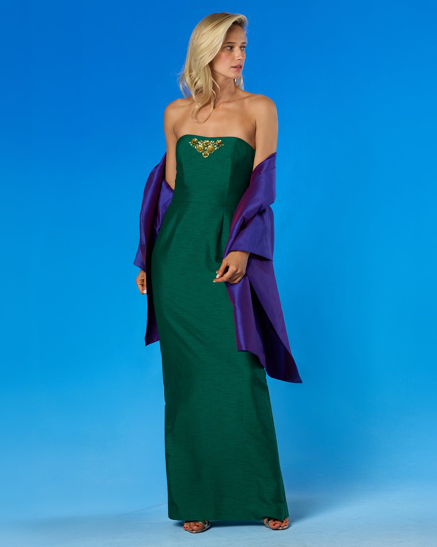 A woman models a strapless emerald green gown with a gold embroidered bodice, styled with a draped purple shawl, set against a solid blue studio background.