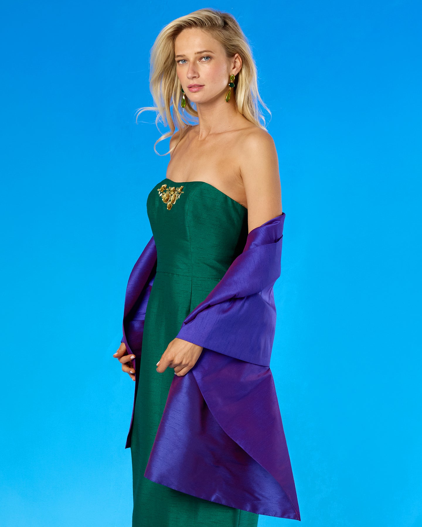 A woman modeling a strapless emerald green dress with a purple draped overlay and a gold embellished bodice, set against a solid blue studio background.