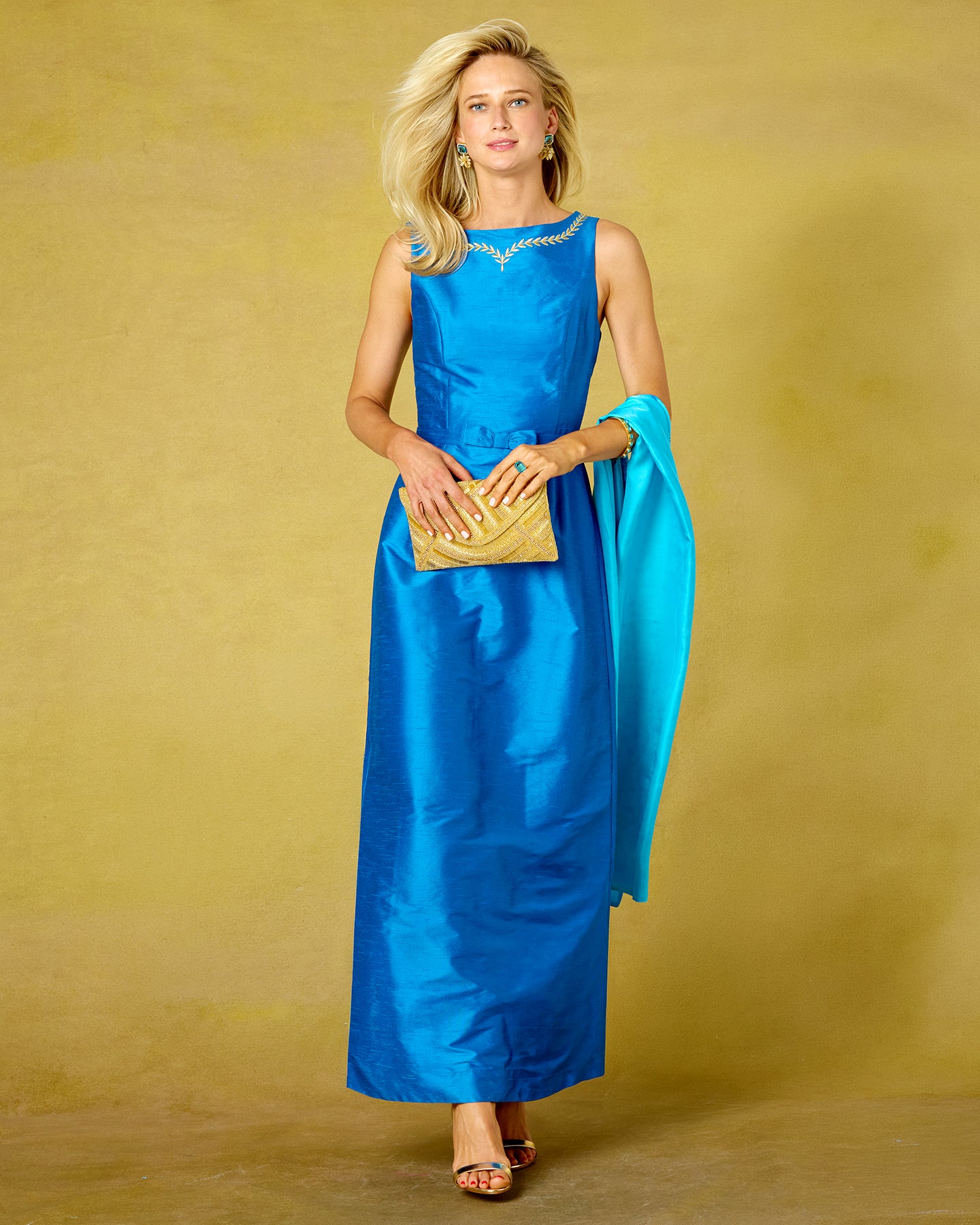A woman modeling a sleeveless bright blue satin maxi dress with gold trim at the neckline, holding a gold clutch and wearing a matching turquoise blue scarf against a textured mustard yellow studio backdrop.