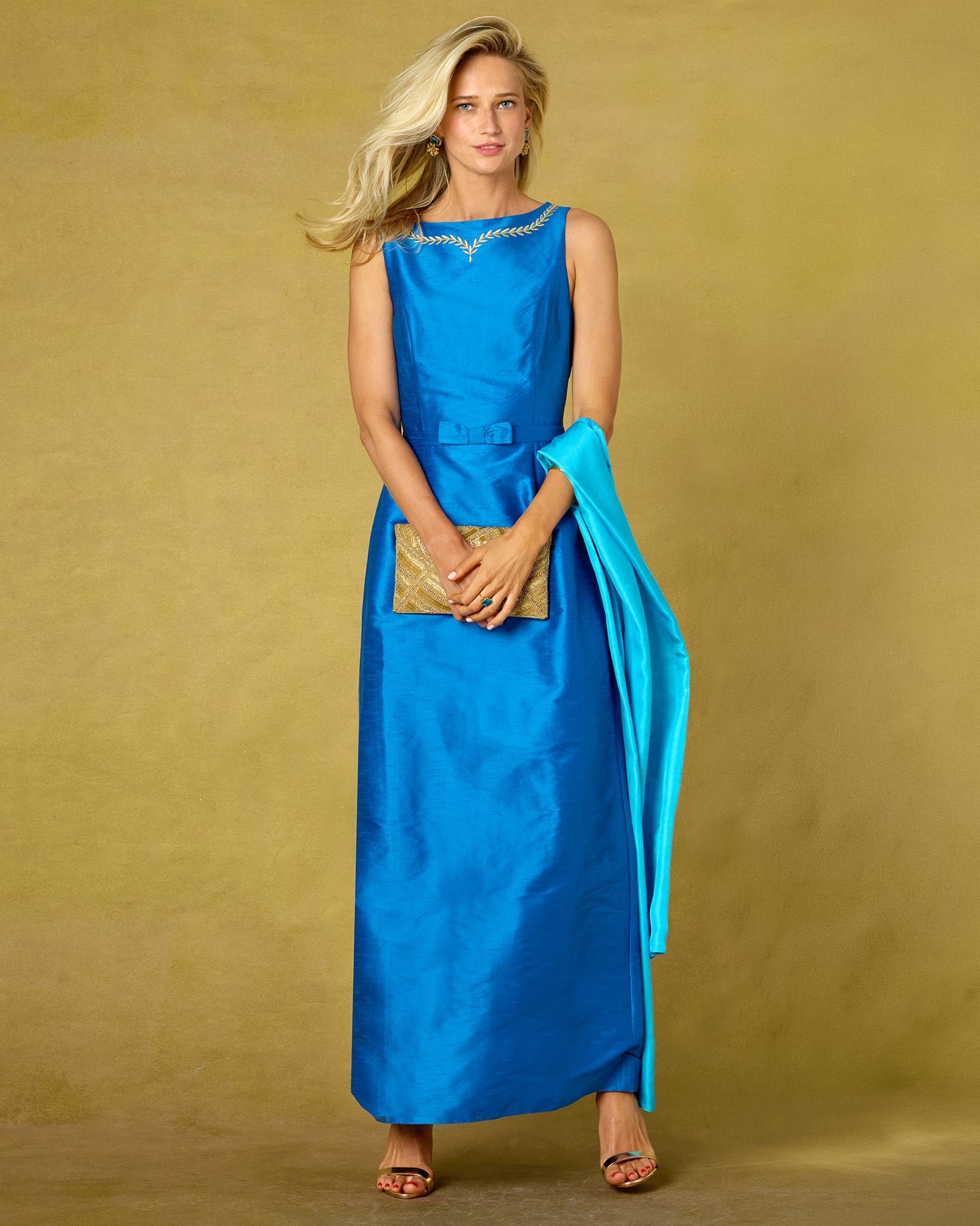 A woman modeling a sleeveless bright blue satin maxi dress with a matching draped scarf and a small gold clutch, set against a textured mustard yellow studio backdrop.