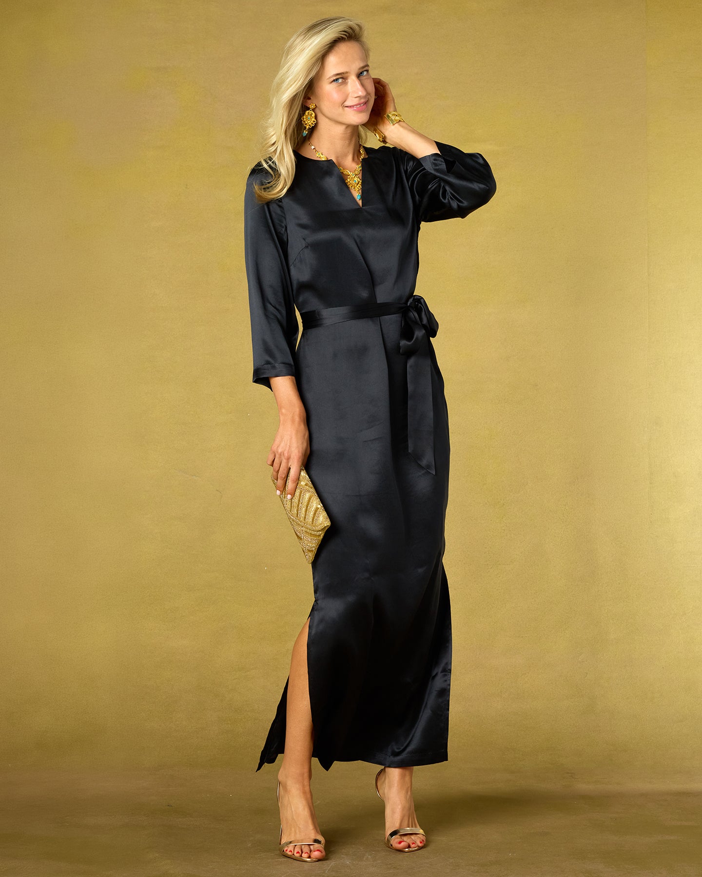 Woman wearing a black satin dress with a gold clutch against a beige background