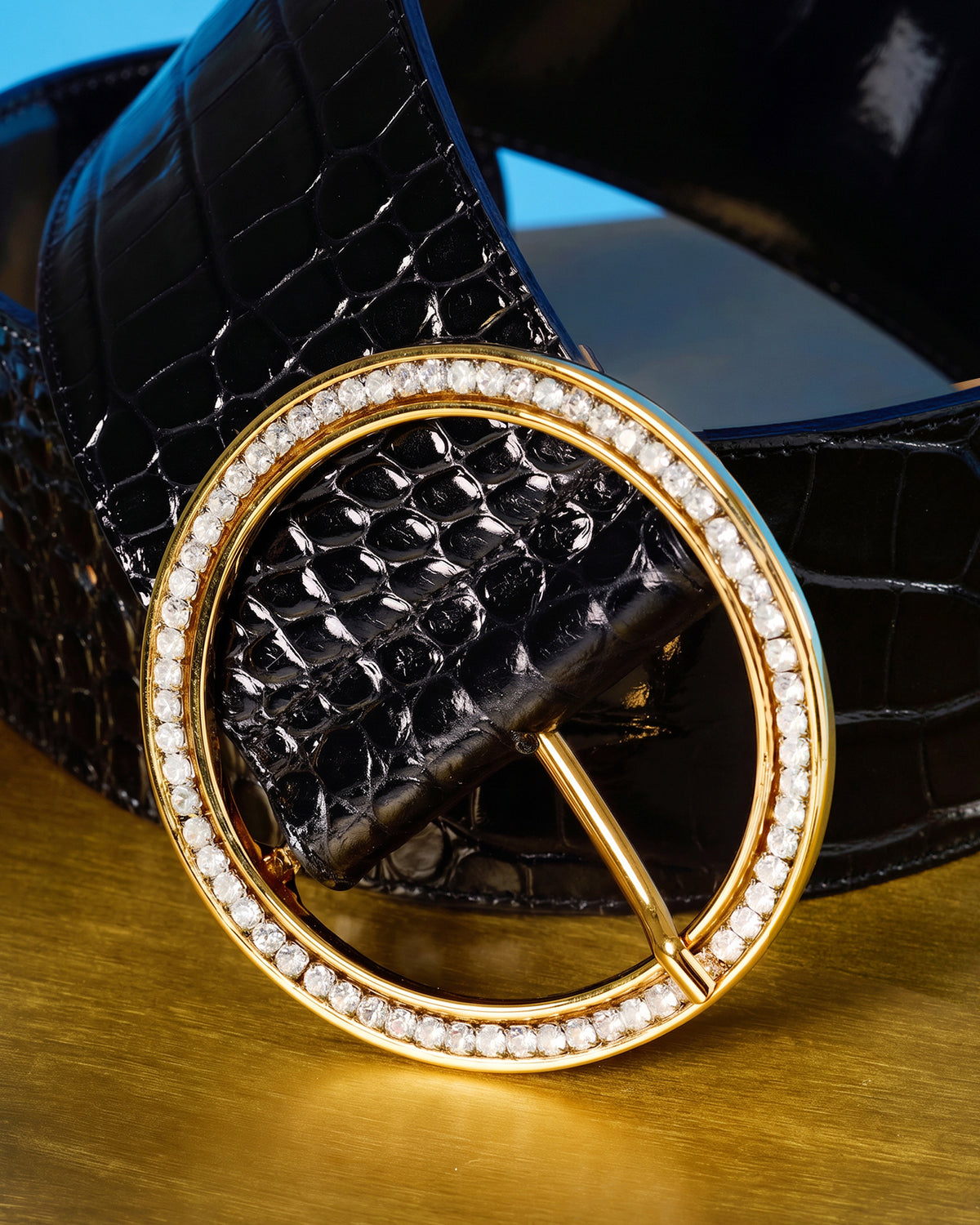A close-up of a black crocodile-embossed belt with a large oval gold-tone buckle encrusted with clear rhinestones, resting on a gold surface.