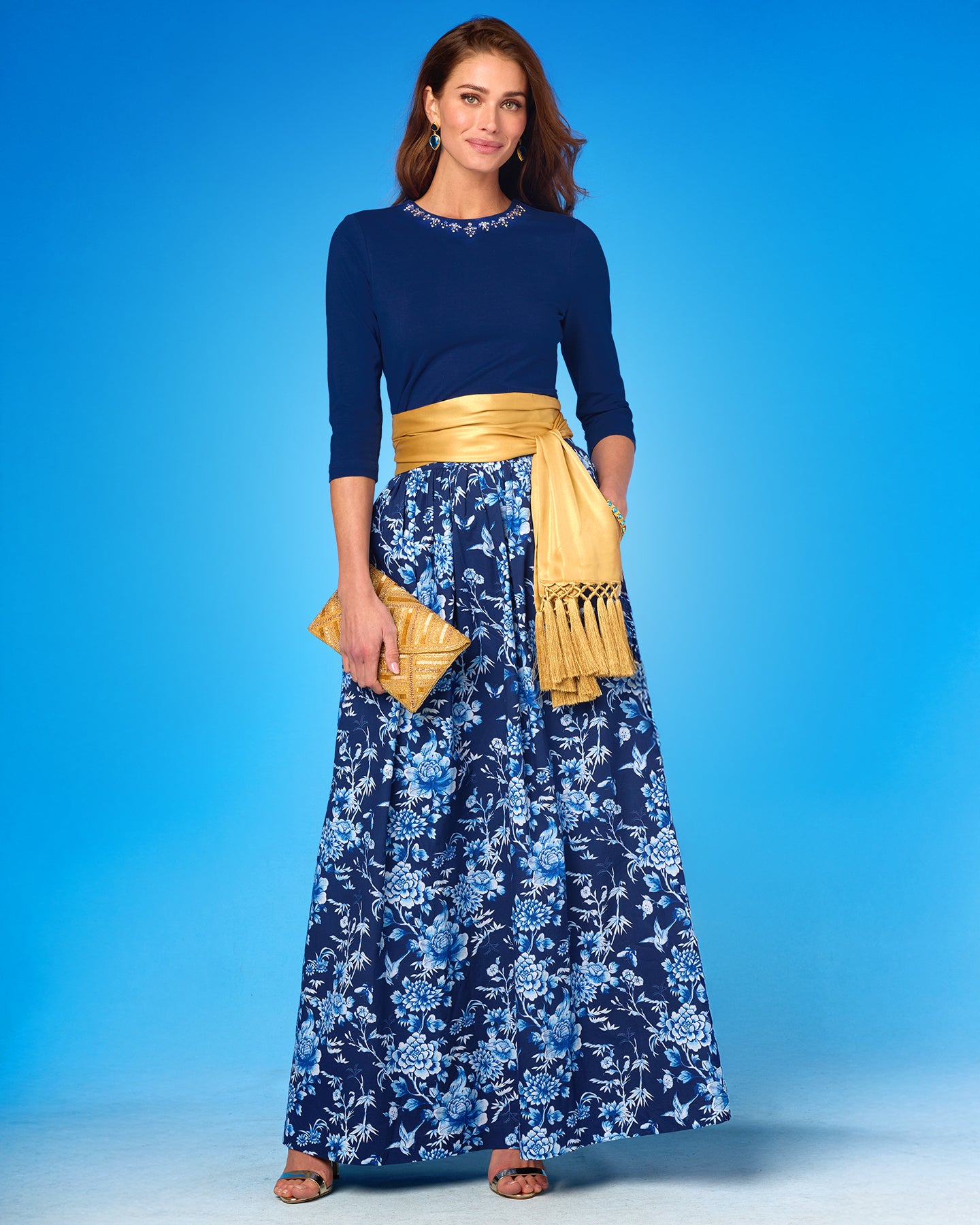 Woman wearing a navy top and floral skirt with a yellow belt and clutch against a blue background