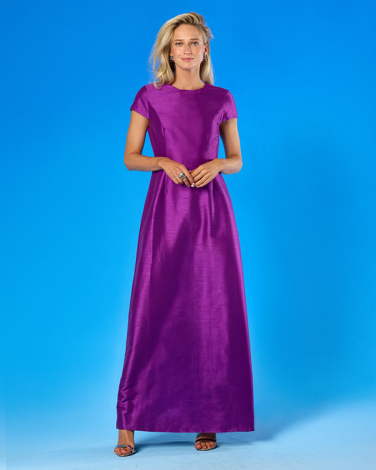 A woman modeling a floor-length purple dress with short sleeves and a round neckline, standing against a solid blue studio background.