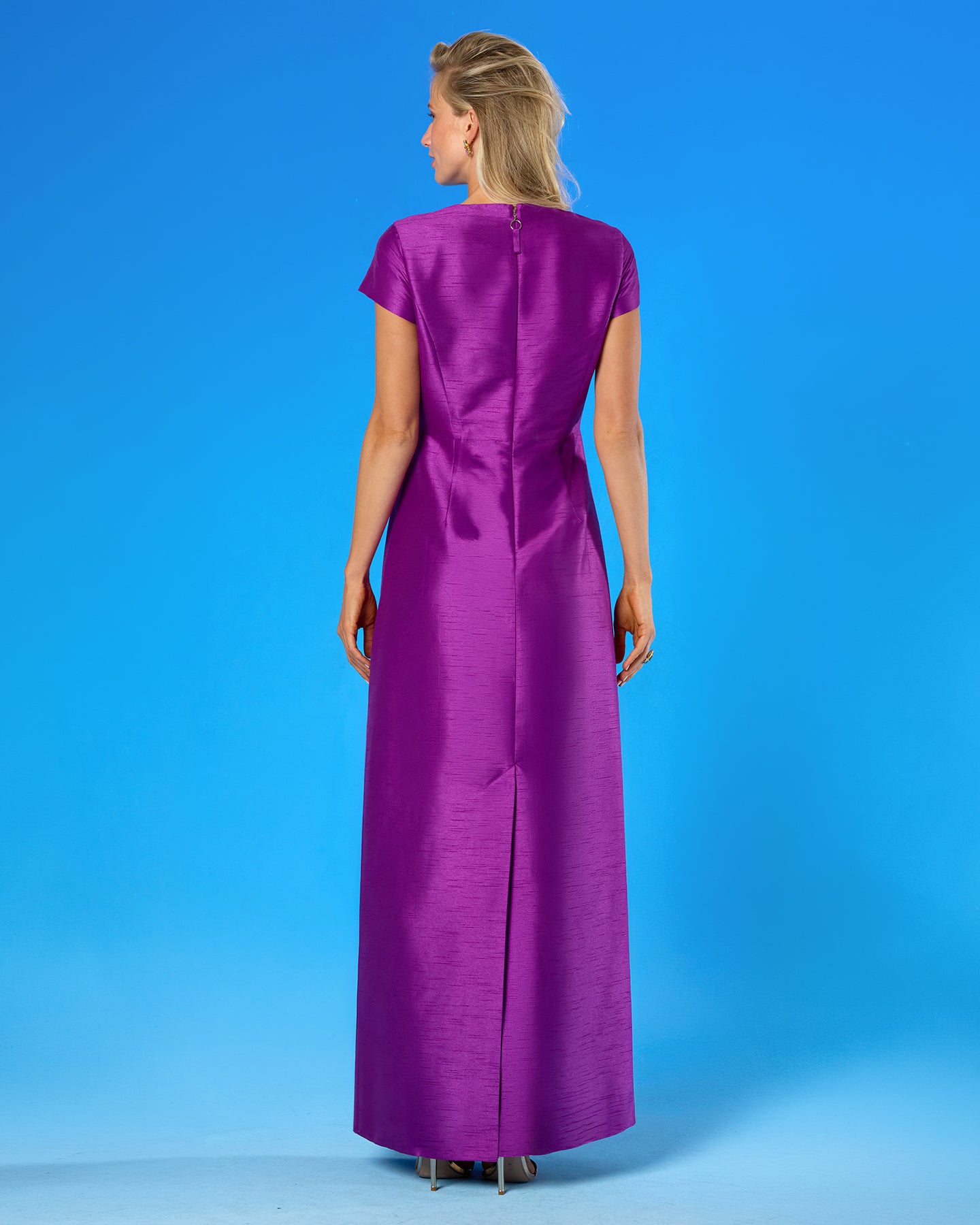 A woman viewed from the back wearing a floor-length magenta-purple dress with short cap sleeves and a front slit, standing against a bright blue studio background.