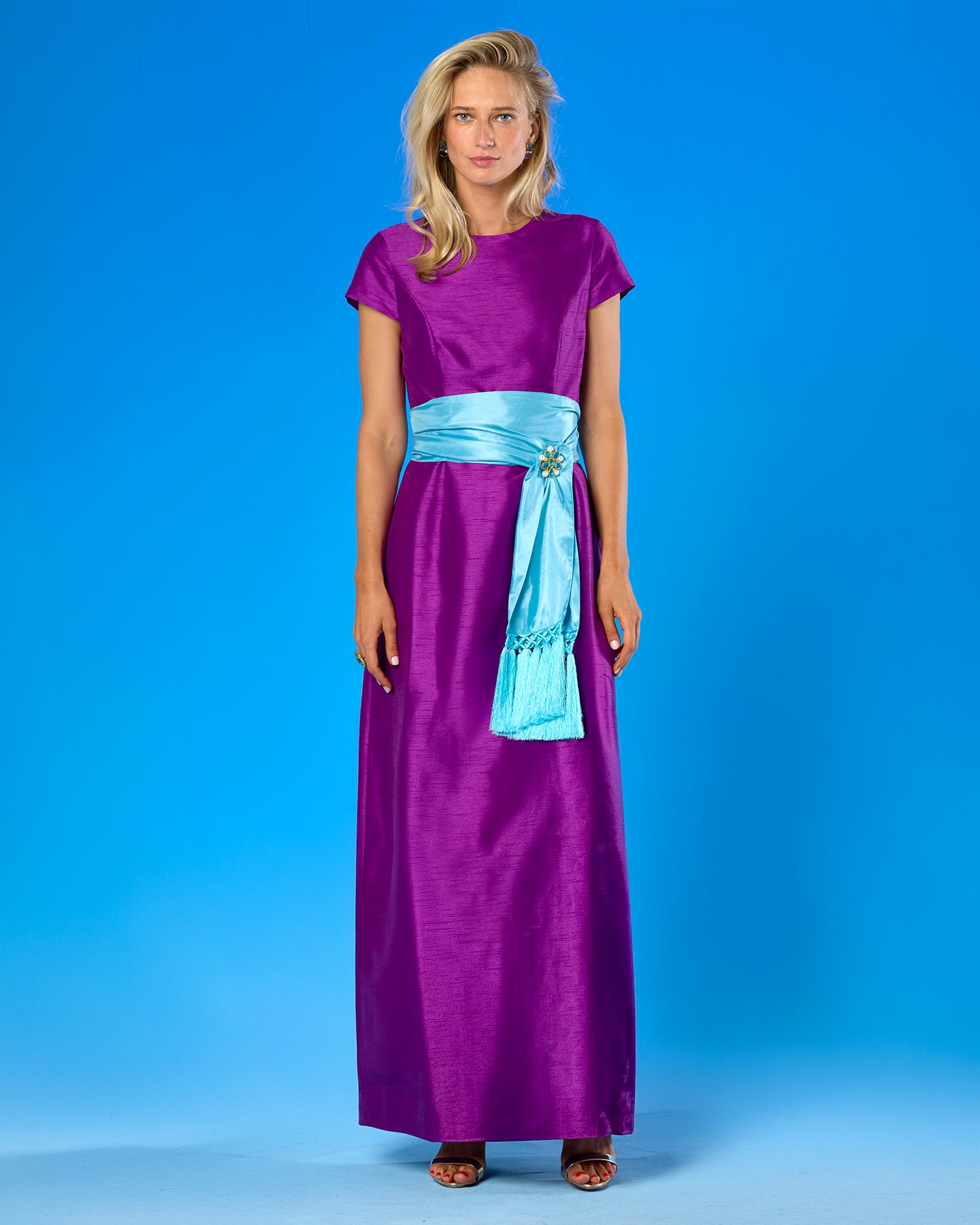 A woman modeling a floor-length purple dress with short cap sleeves and a bright turquoise sash at the waist, standing against a solid blue studio background.