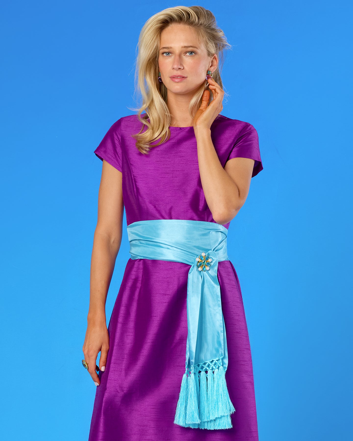 A woman modeling a purple short-sleeve dress with a light blue sash at the waist, posed against a solid blue studio background.