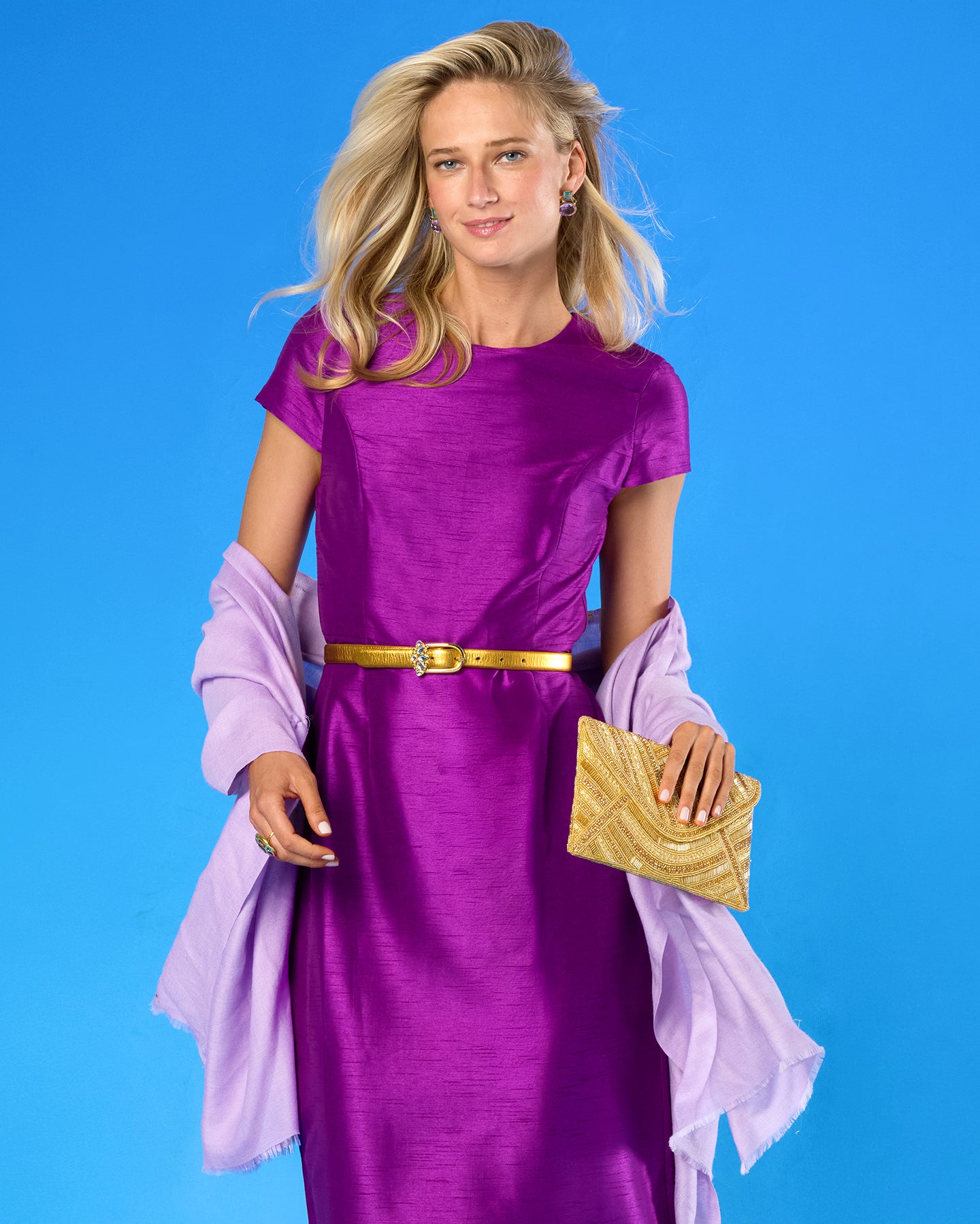 A woman modeling a short-sleeve purple dress with a gold belt, holding a gold clutch and draped in a light purple shawl against a solid blue background.
