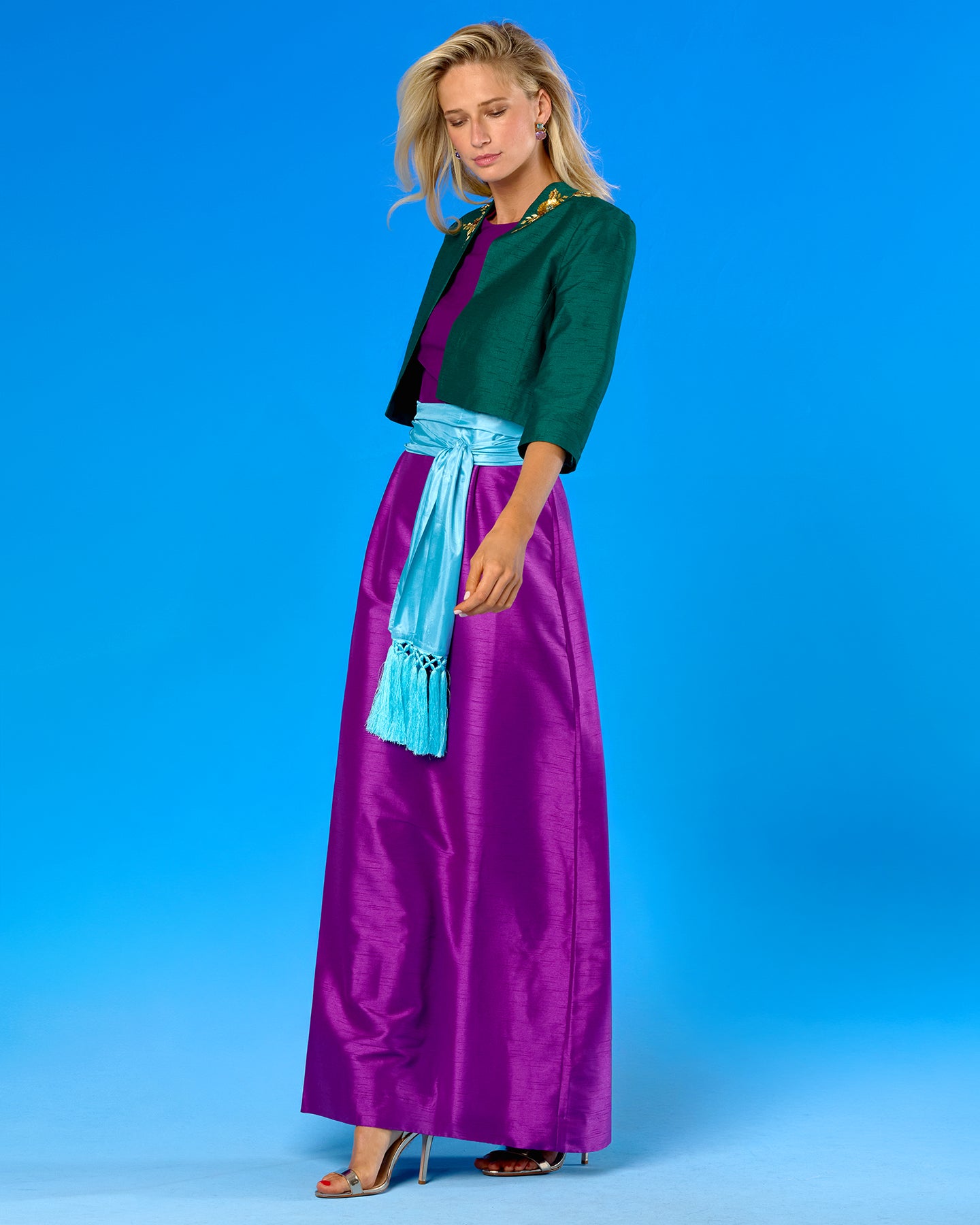 A woman modeling a long purple satin skirt with a light blue sash, paired with a dark green cropped jacket, against a solid blue studio background.
