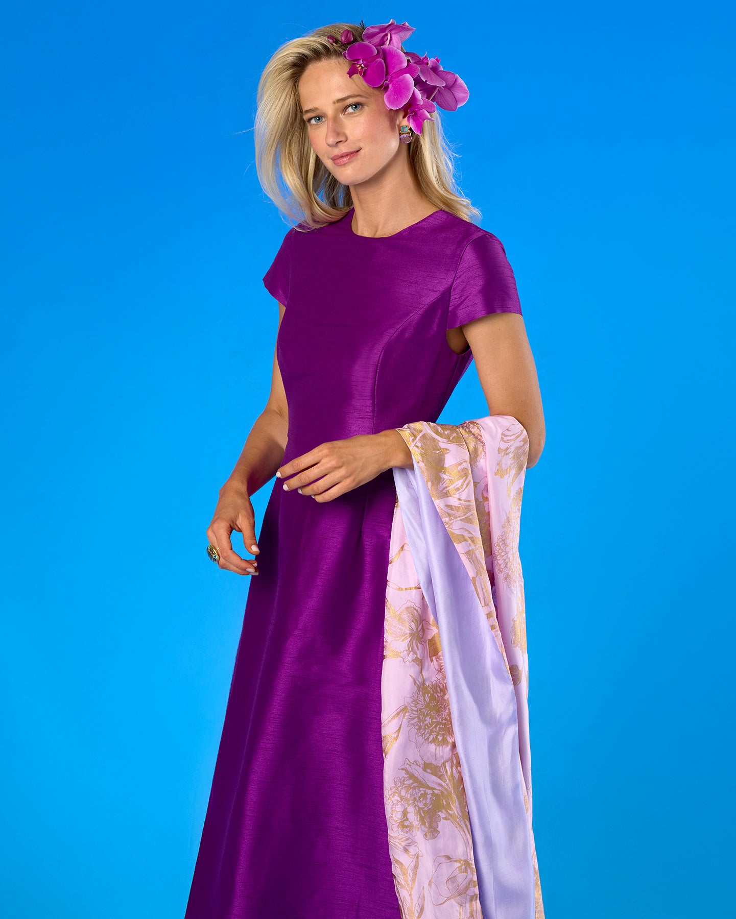 A woman modeling a purple satin maxi dress with a light purple and gold patterned dupatta, set against a solid blue studio background.