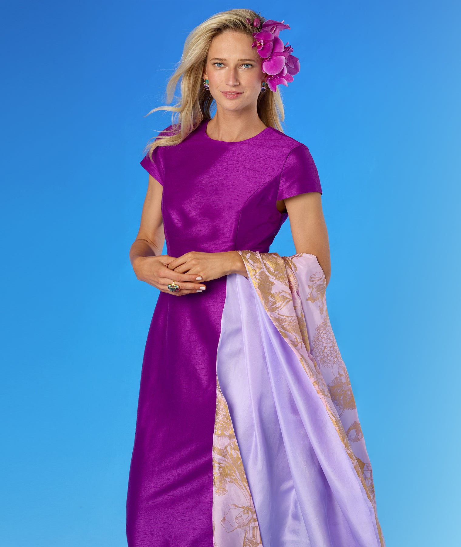 A woman modeling a short-sleeved magenta dress with a light purple dupatta featuring a gold botanical pattern, set against a solid blue studio background.