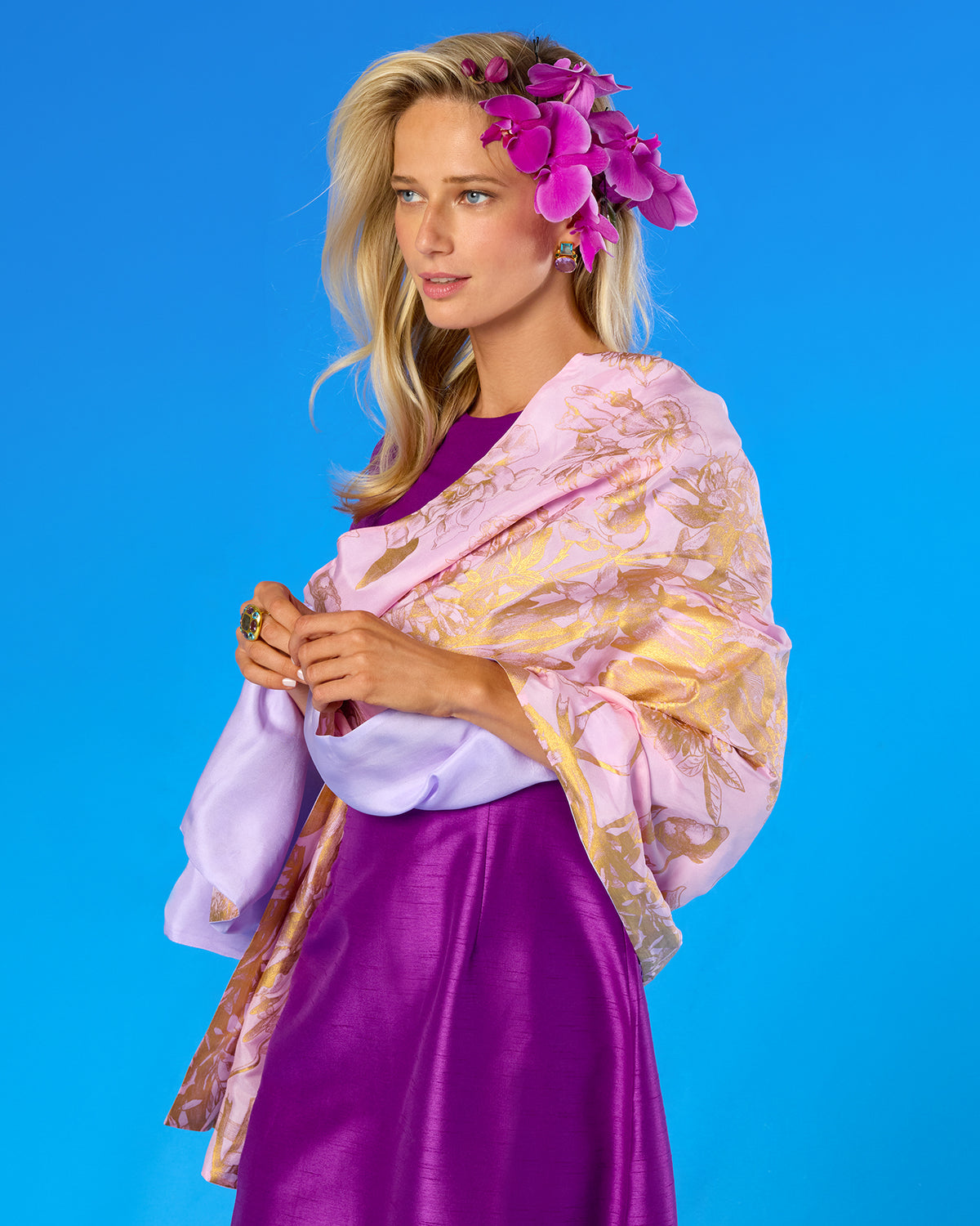 A woman modeling a light pink and gold patterned shawl draped over a purple dress, with a matching purple orchid flower crown, set against a solid blue studio background.