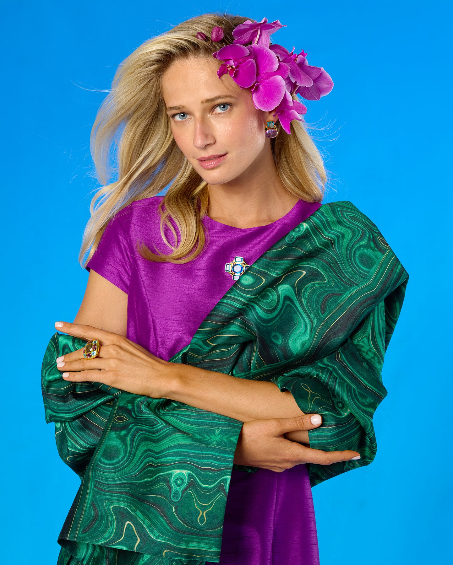 A blonde model wearing a purple short-sleeve top and a green patterned shawl draped over her shoulders, with pink orchid flowers in her hair, posed against a bright blue studio background.