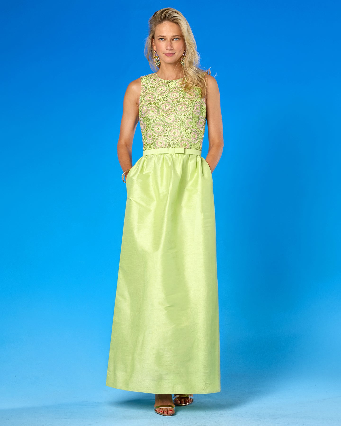 A woman modeling a sleeveless lime green maxi dress with a floral embroidered bodice and a satin skirt, standing against a solid blue studio background.