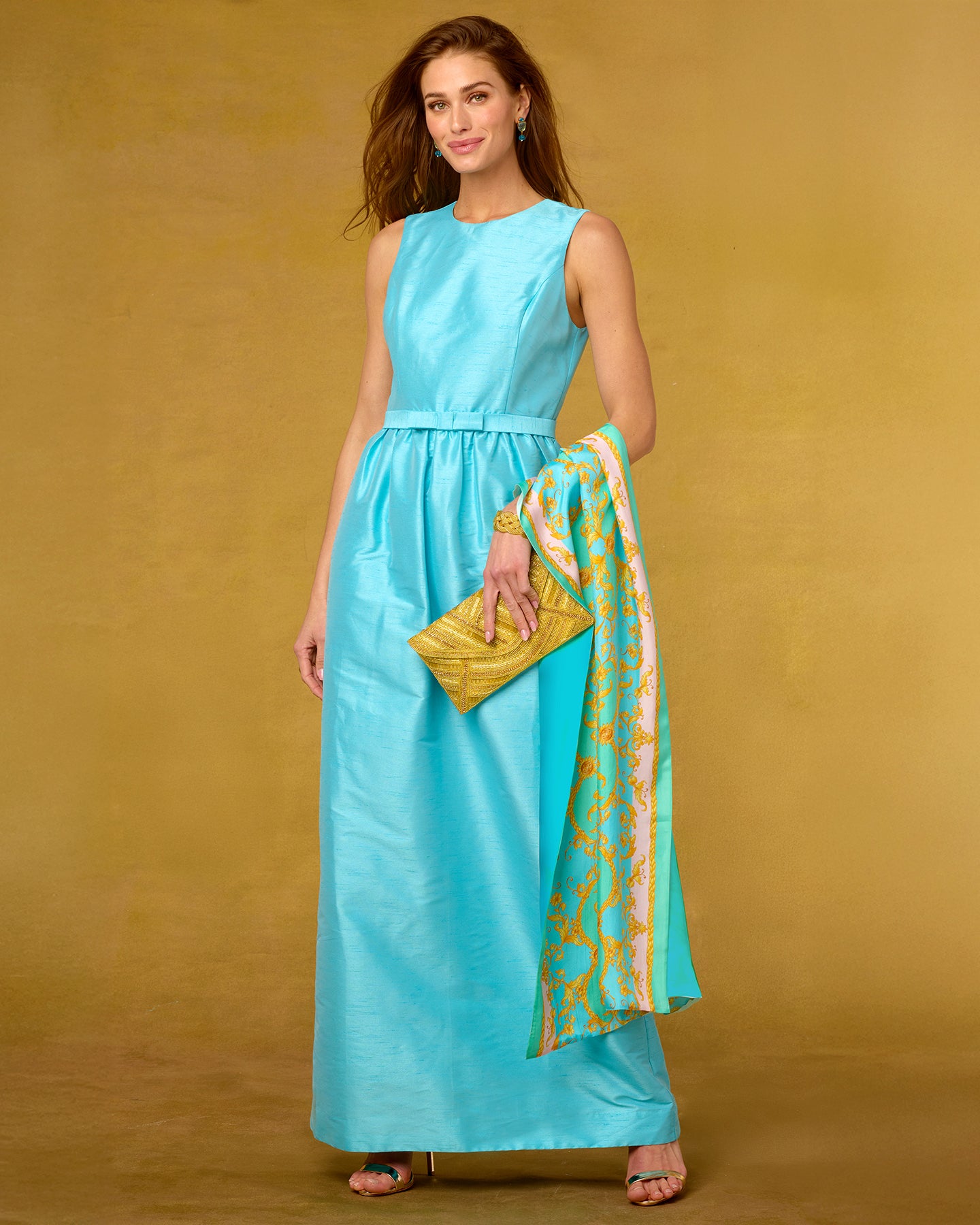 Woman in a turquoise dress holding a yellow clutch and patterned scarf against a beige background