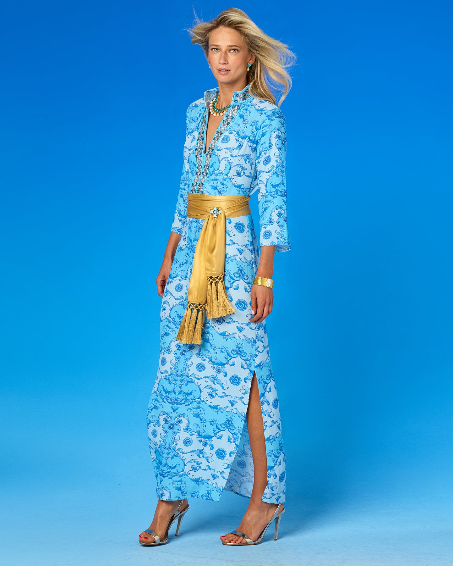 A woman modeling a light blue and white floral maxi dress with a wide gold sash belt, standing against a solid blue studio background.