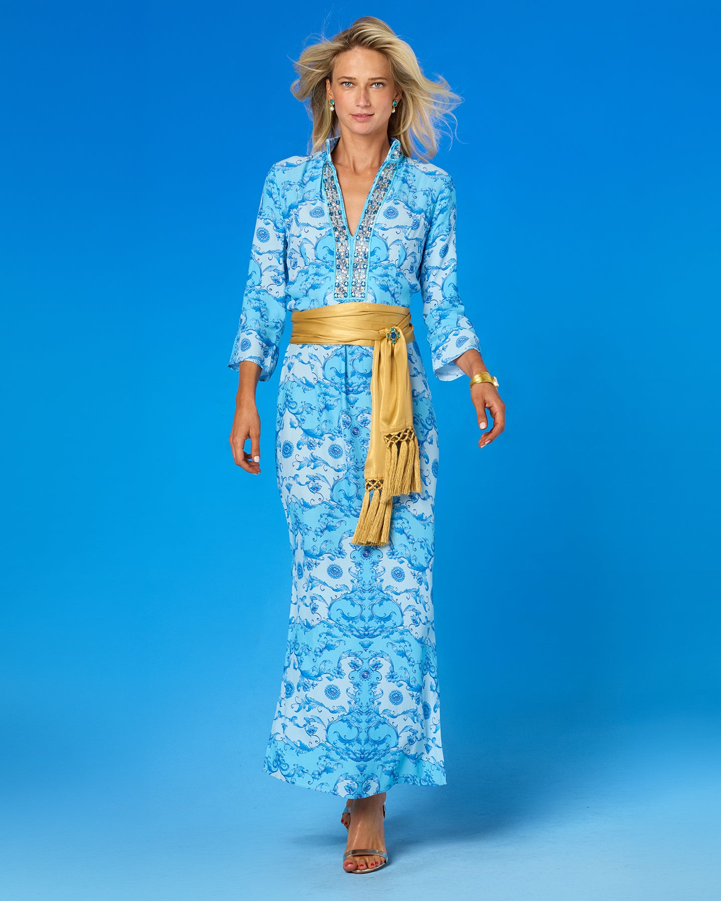 A woman models a light blue maxi dress with a white floral pattern and a wide gold belt, standing against a solid blue studio background.