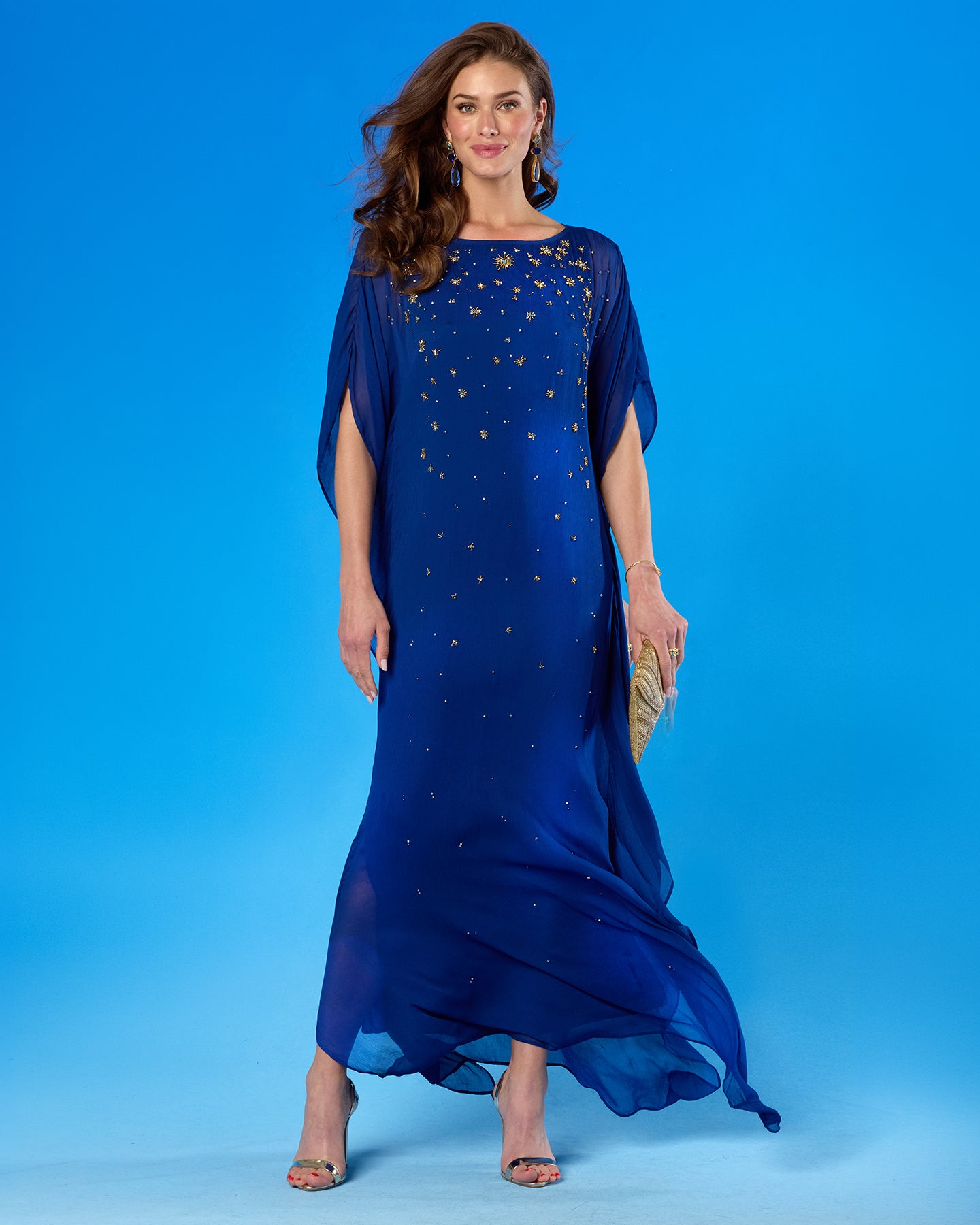 A woman modeling a flowing royal blue maxi dress with gold star-like embellishments, standing against a solid blue studio background.