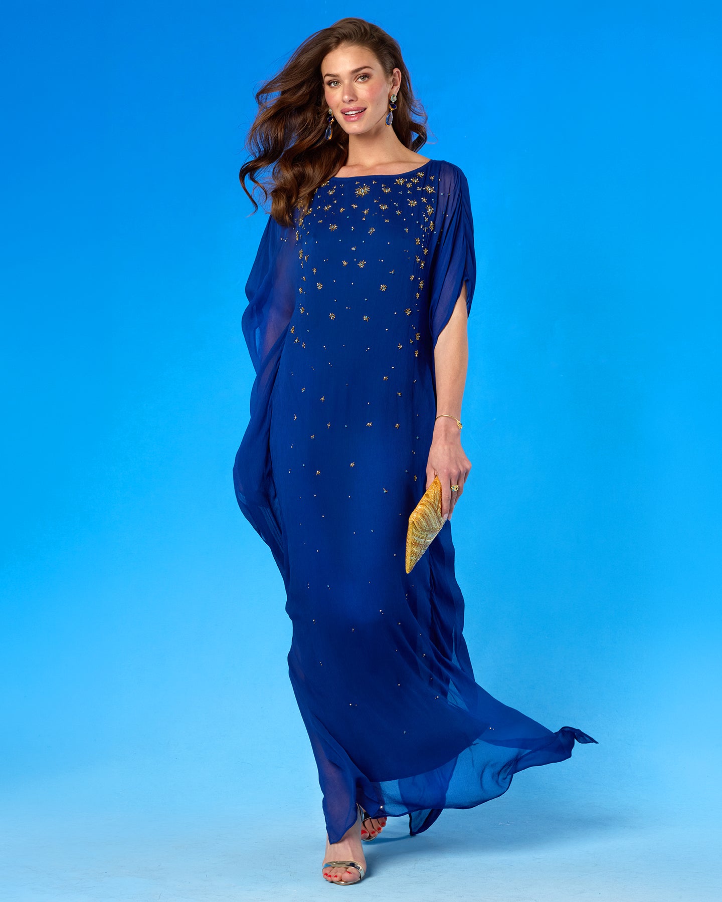 A woman modeling a flowing royal blue maxi dress with gold star-like embellishments, standing against a solid blue studio background and holding a gold clutch.