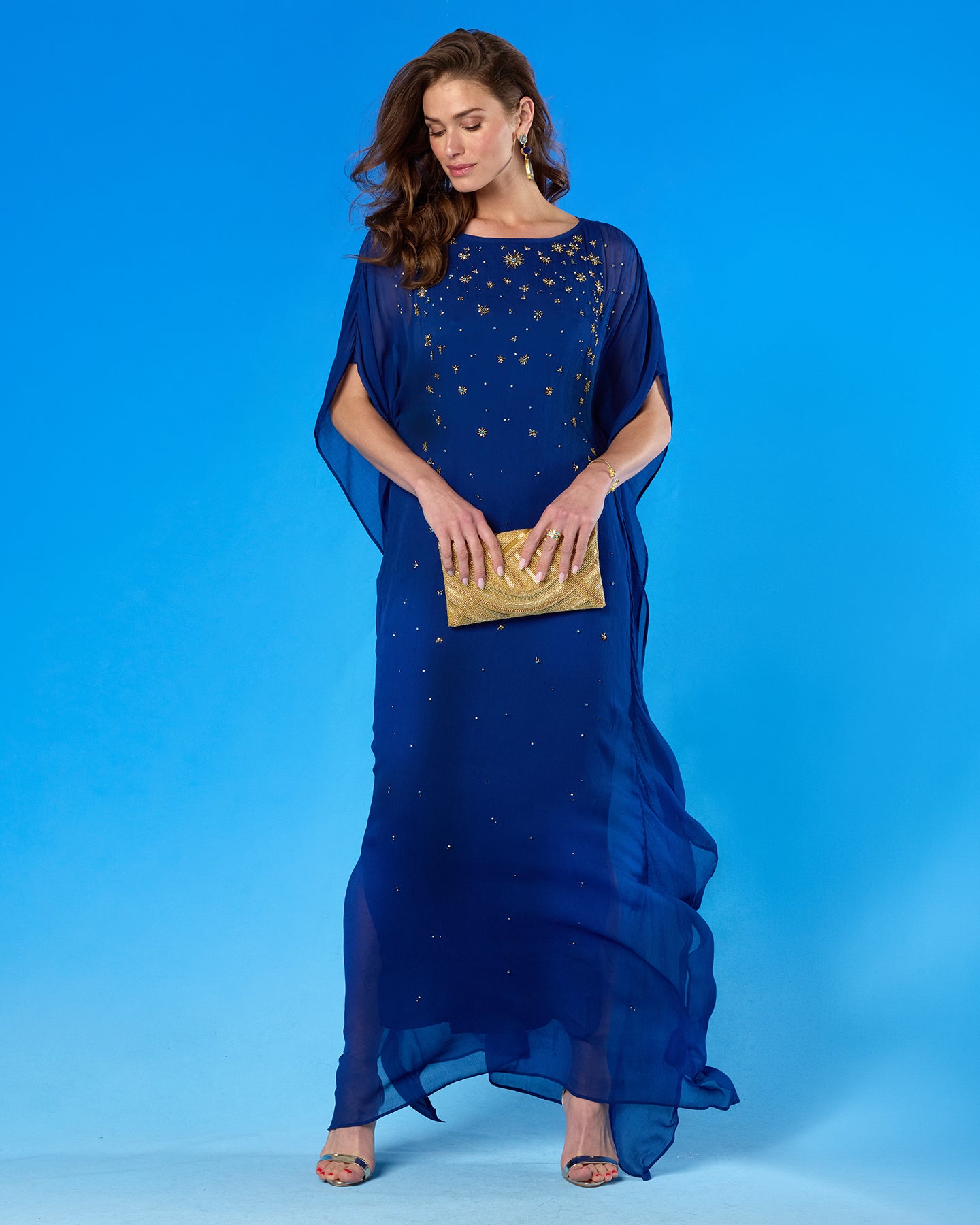 A woman modeling a floor-length royal blue sheer gown with scattered gold star embellishments, holding a metallic gold clutch against a solid blue studio background.