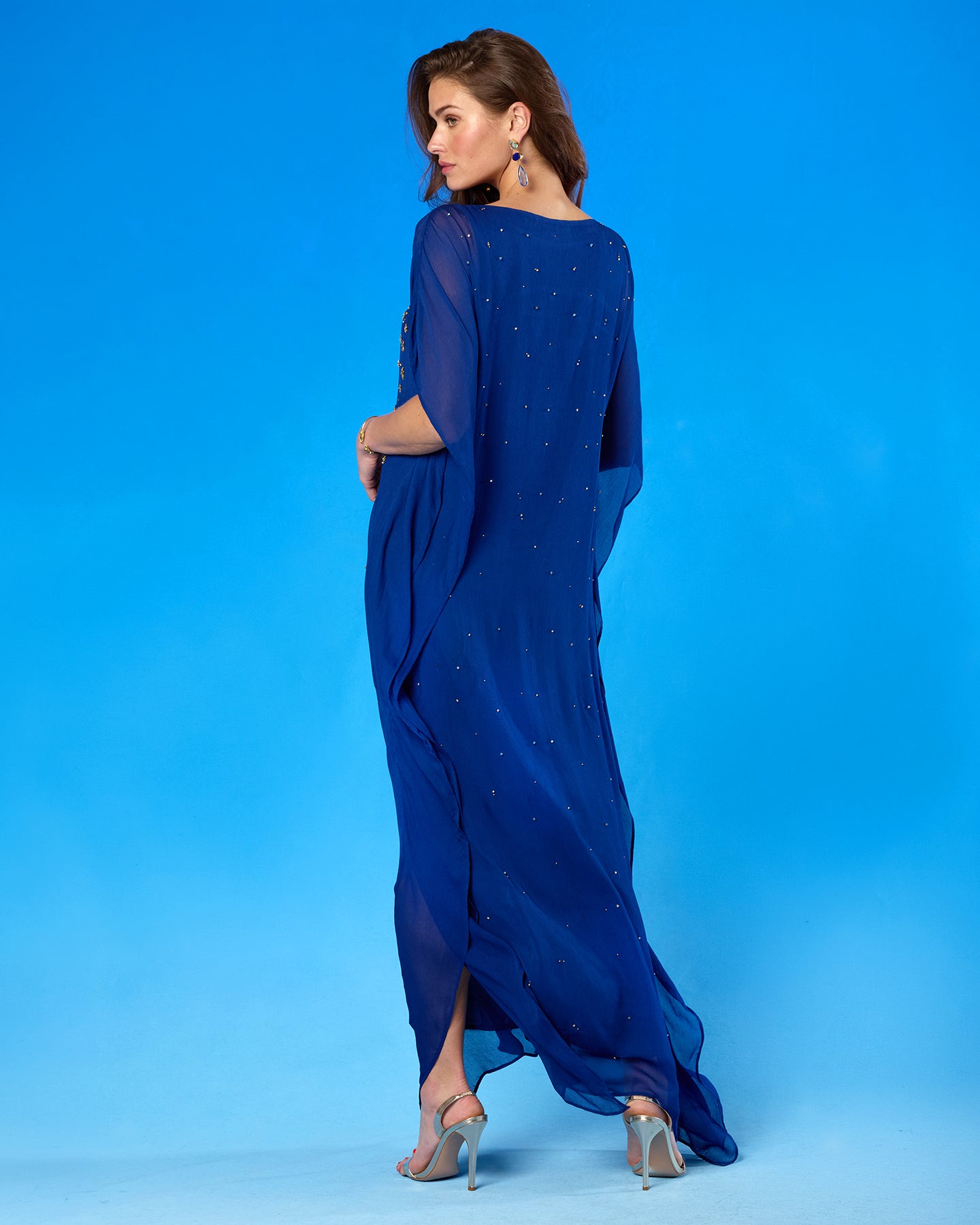 A woman modeling a royal blue sheer maxi dress with subtle sequin embellishments, shown from a side-back angle against a solid blue studio background.