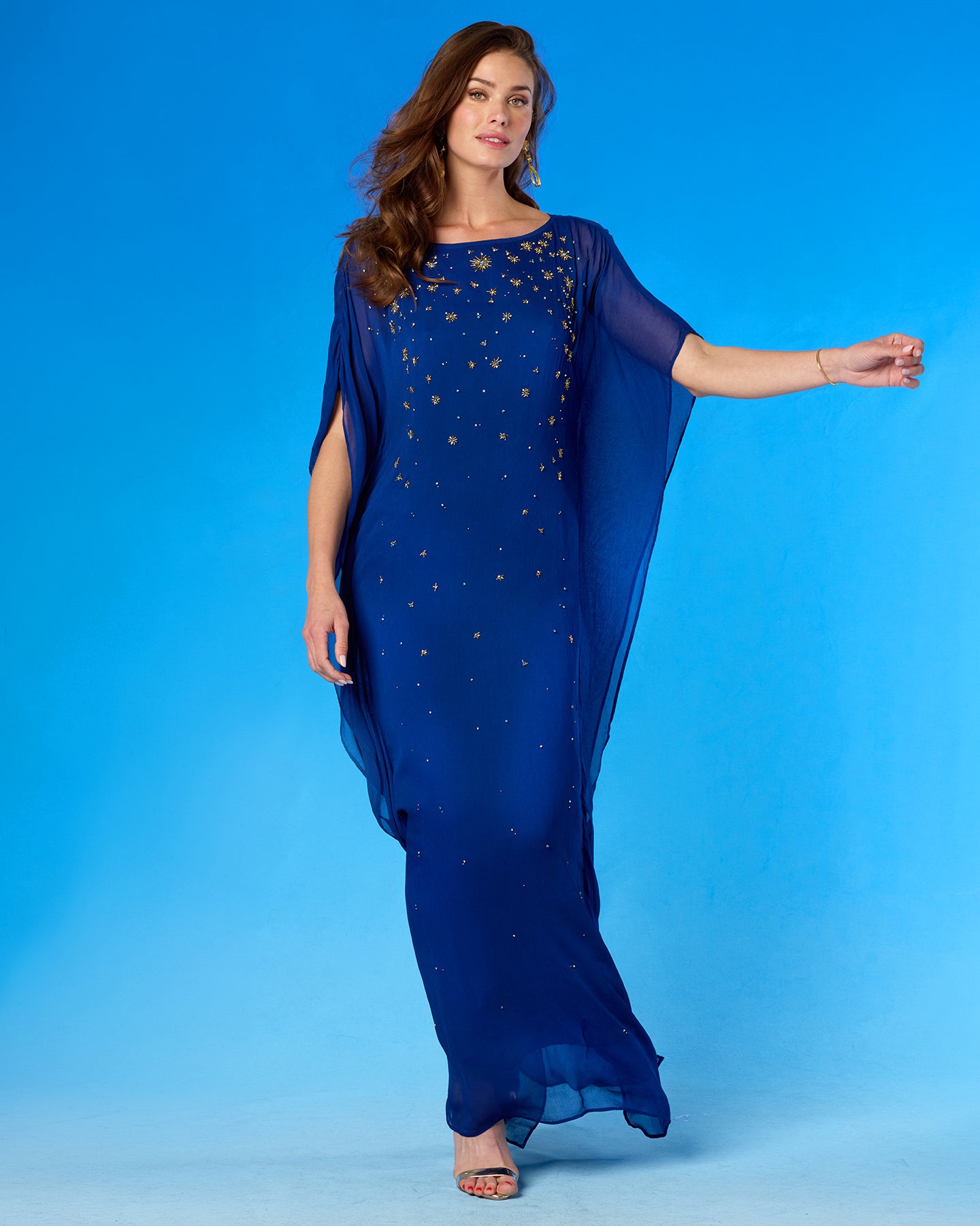 A woman modeling a royal blue maxi dress with sheer draped cap sleeves and scattered gold sequin embellishments, set against a solid blue studio background.
