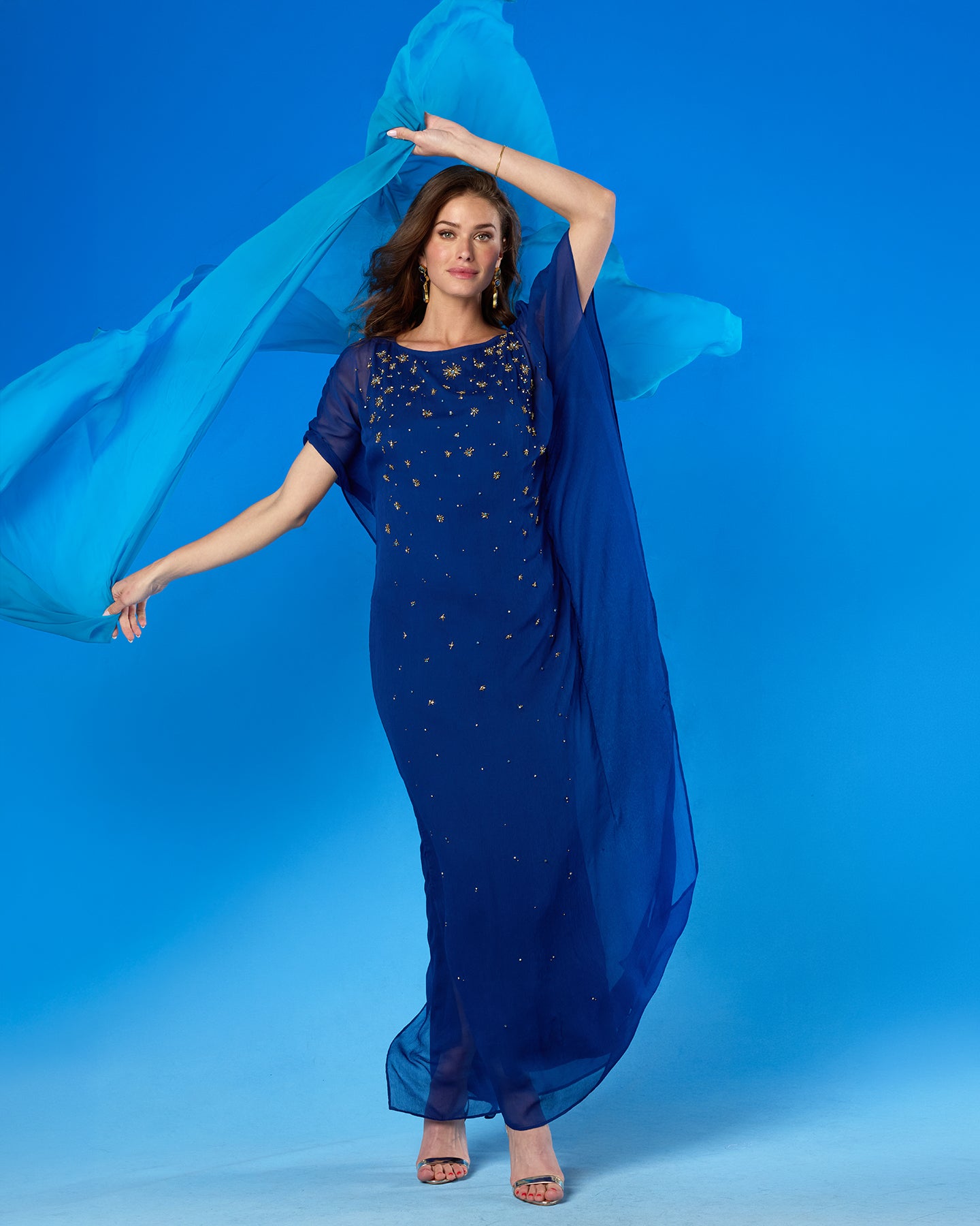A woman modeling a royal blue floor-length gown with sheer sleeves and scattered gold star embellishments, holding a matching flowing sheer blue drape against a solid blue studio background.