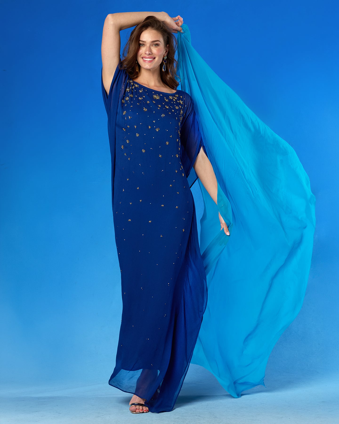 A woman modeling a floor-length navy blue gown with gold star-like embellishments and a flowing turquoise-blue sheer drape, posed against a solid blue studio background.