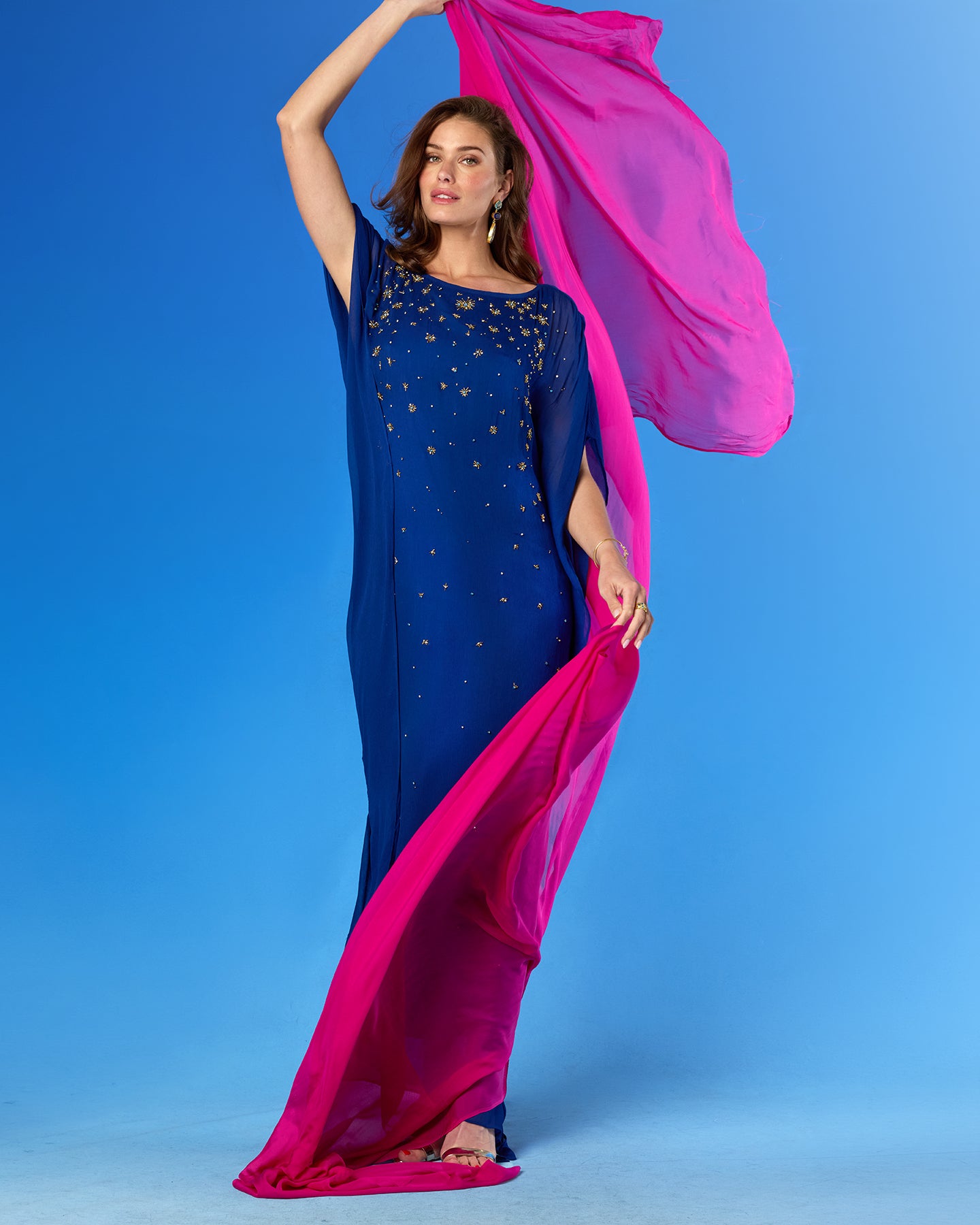 A woman modeling a royal blue sleeveless gown with gold star embroidery and a bright pink dupatta, posed against a solid blue studio background.