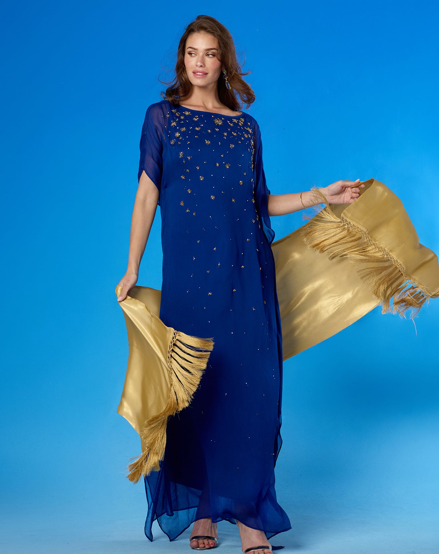 A woman modeling a royal blue floor-length dress with gold embroidered details and a flowing gold dupatta, posed against a solid blue studio background.