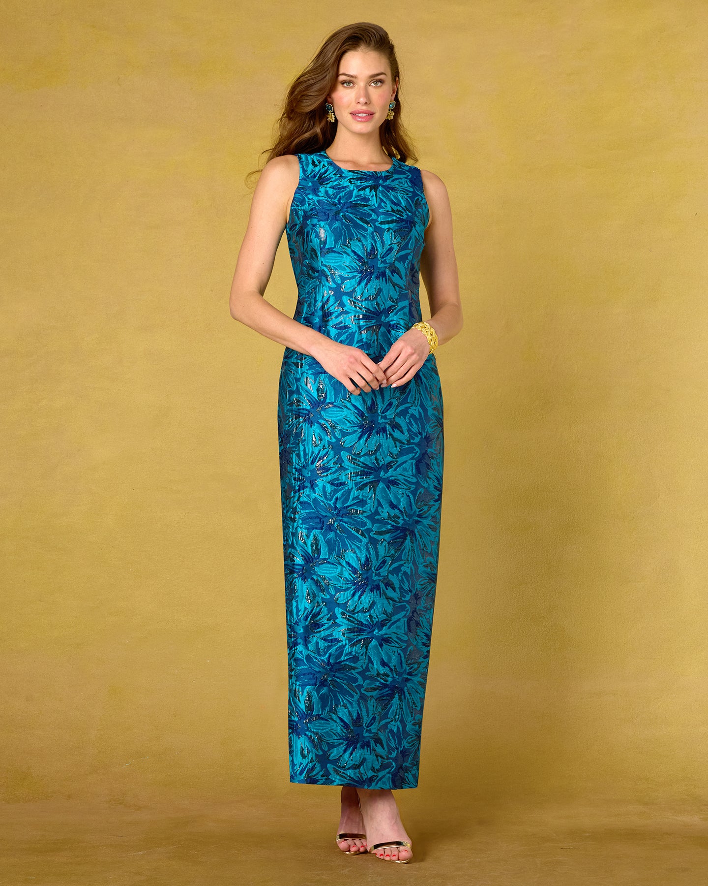 A woman modeling a sleeveless, floor-length teal dress with a dark blue tropical floral pattern, standing against a textured mustard yellow studio backdrop.