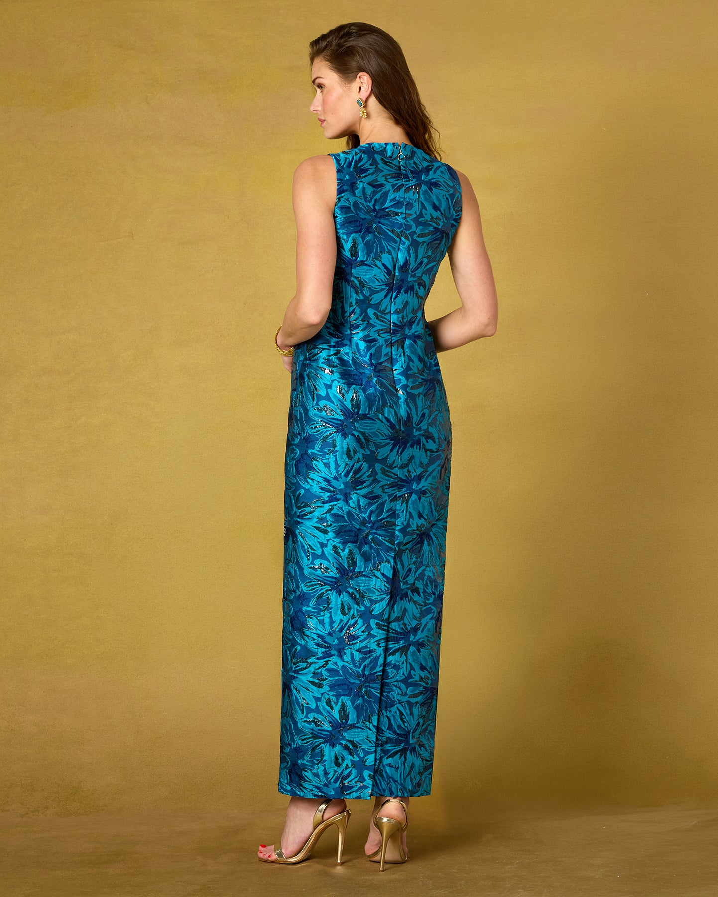 A woman modeling a sleeveless teal and blue floral maxi dress from the back, standing against a textured mustard yellow studio backdrop.