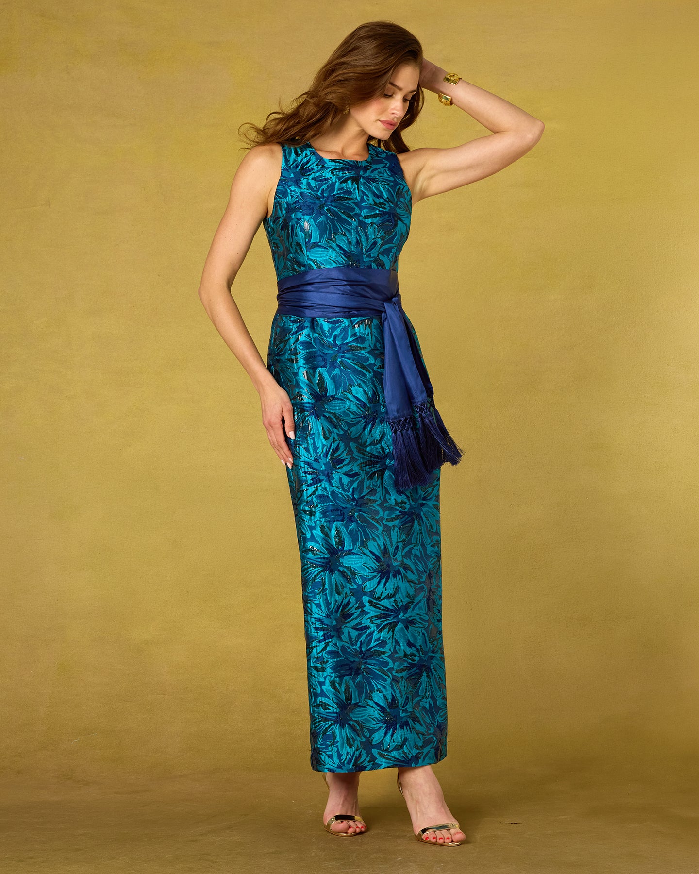 A woman modeling a sleeveless maxi dress with a teal and blue tropical floral pattern and a dark blue satin waist sash, posed against a solid mustard yellow studio backdrop.