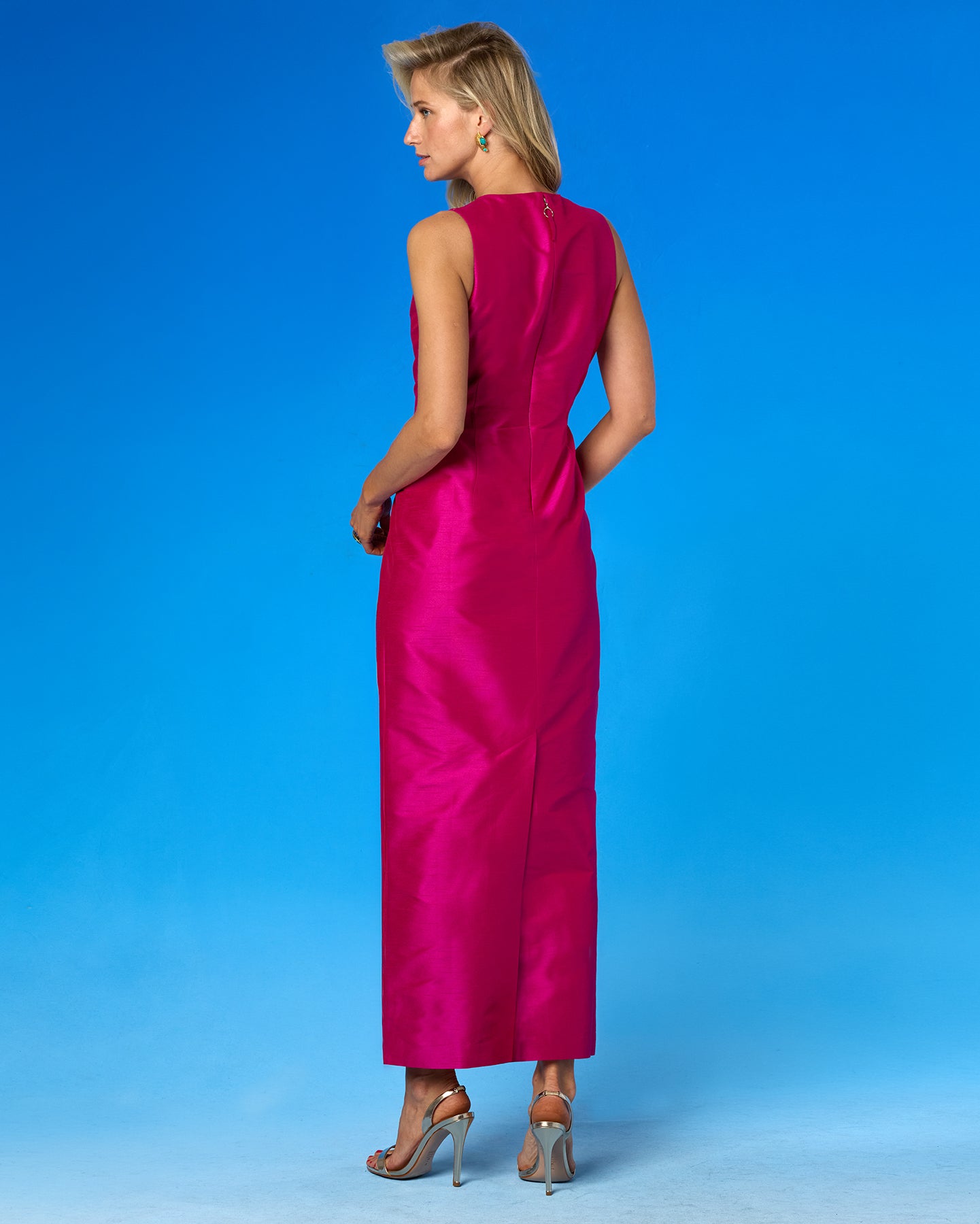 A woman modeling a sleeveless magenta maxi dress from the back, standing against a solid blue studio background.