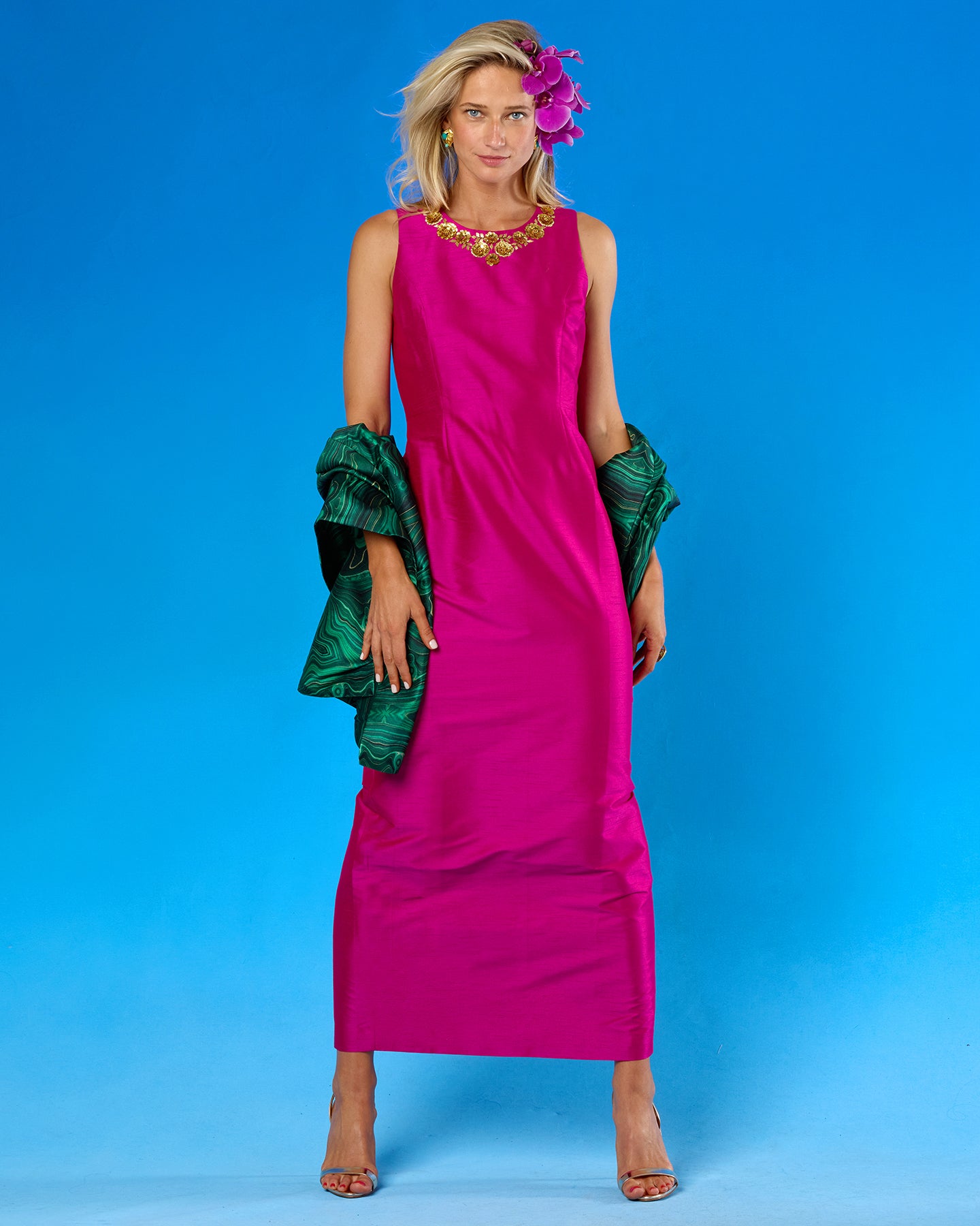 A woman modeling a sleeveless magenta maxi dress with a gold embellished neckline, holding a green patterned wrap, set against a bright blue studio background.