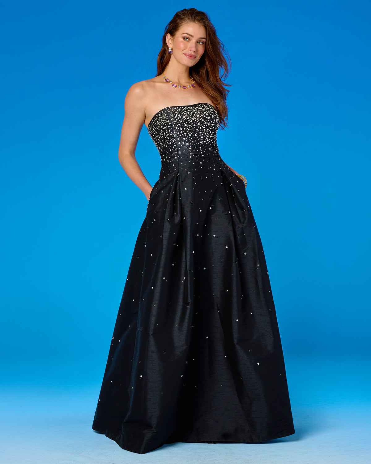 A woman modeling a strapless black ball gown with a heavily embellished beaded bodice and scattered rhinestone-like accents on the full skirt, posed against a bright blue studio backdrop.