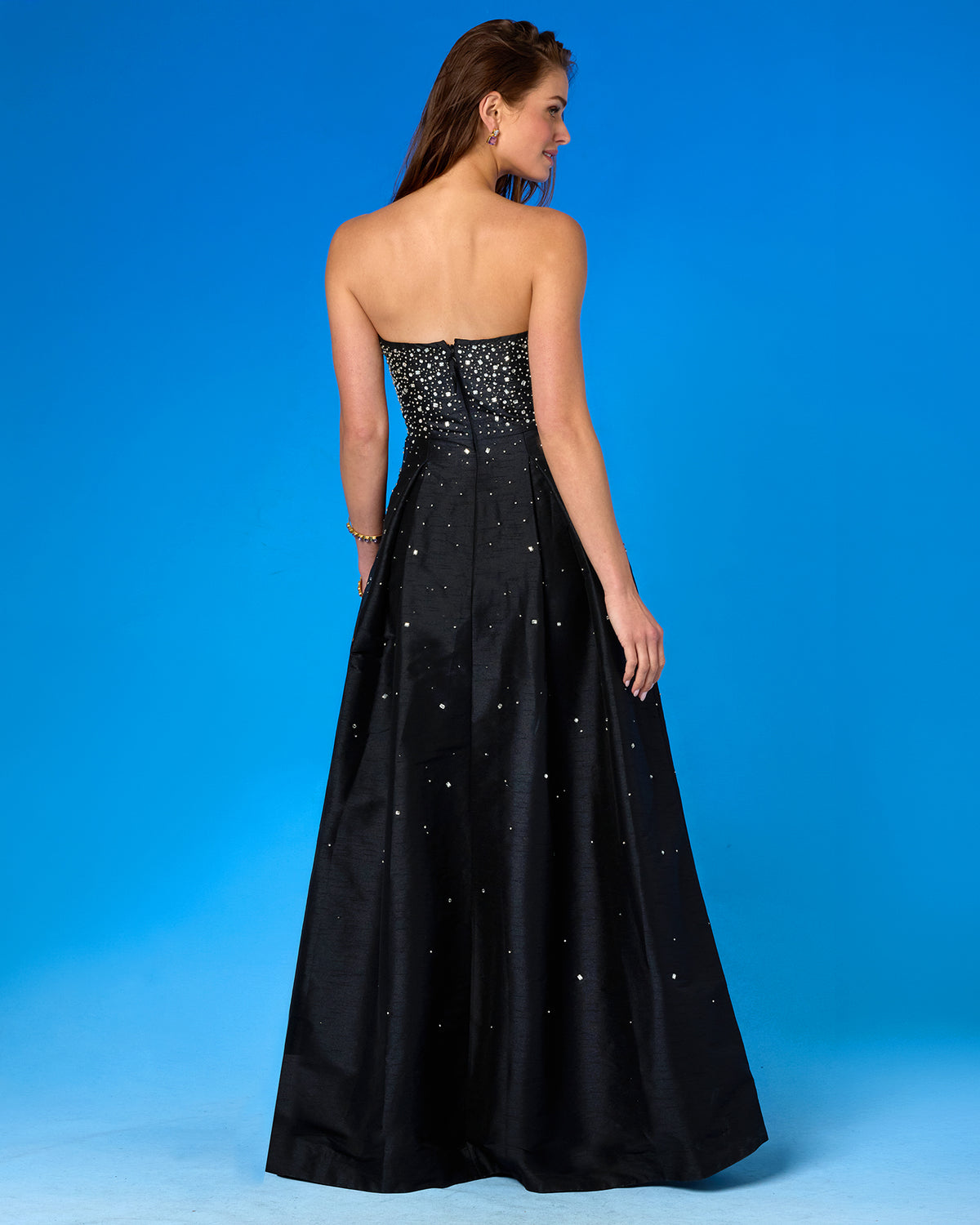 A woman modeling a strapless black formal gown from the back, featuring a beaded bodice and scattered rhinestone embellishments on the full skirt, set against a solid blue studio background.