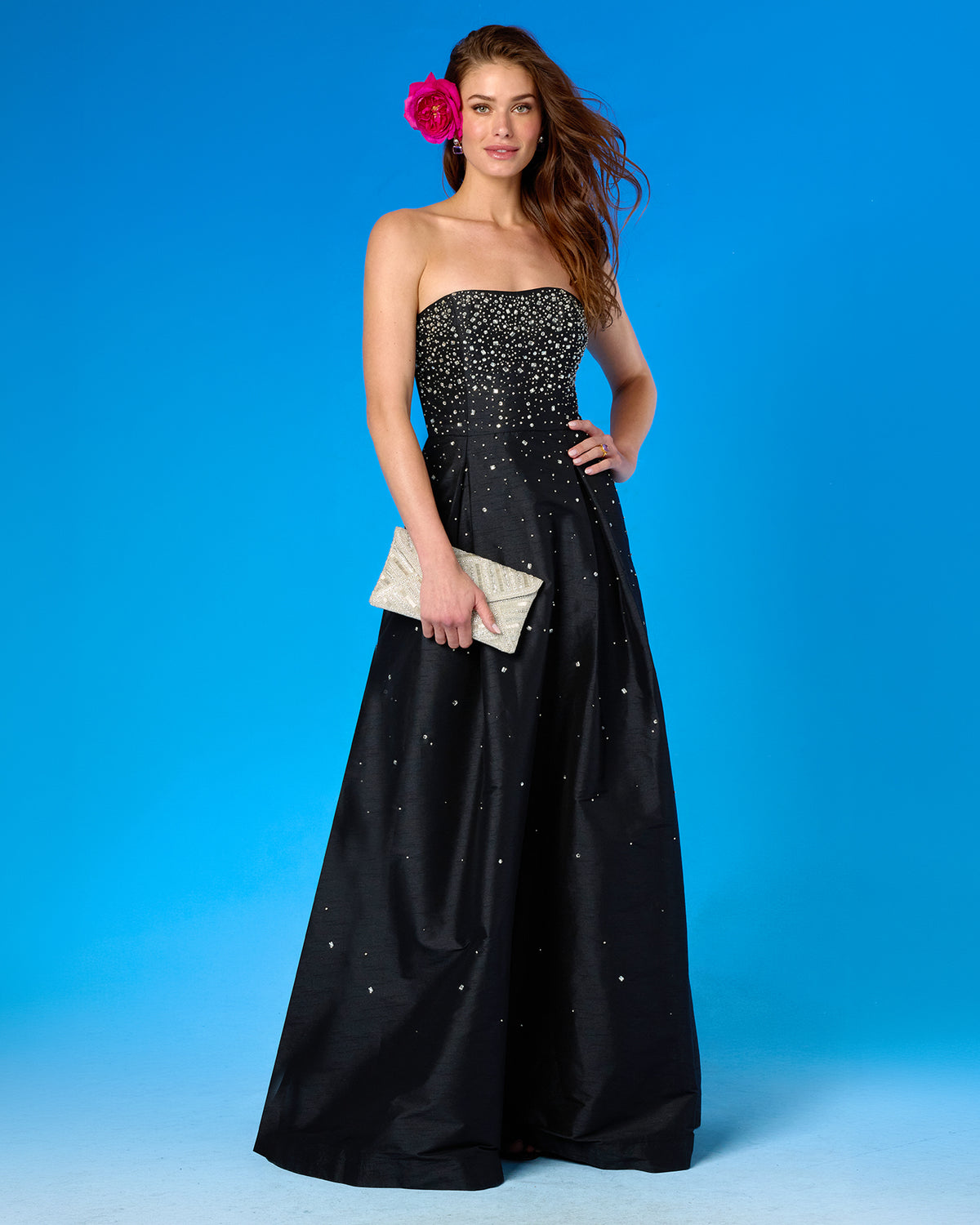 A woman modeling a strapless black taffeta gown with silver rhinestone embellishments, holding a textured cream clutch against a bright blue studio background.