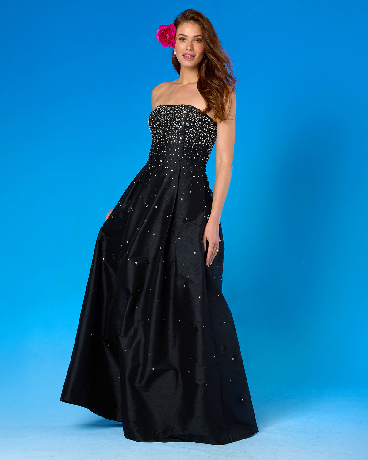 A woman modeling a strapless black taffeta evening gown with a heavily embellished beaded bodice and scattered rhinestone accents on the skirt, posed against a solid blue studio background.