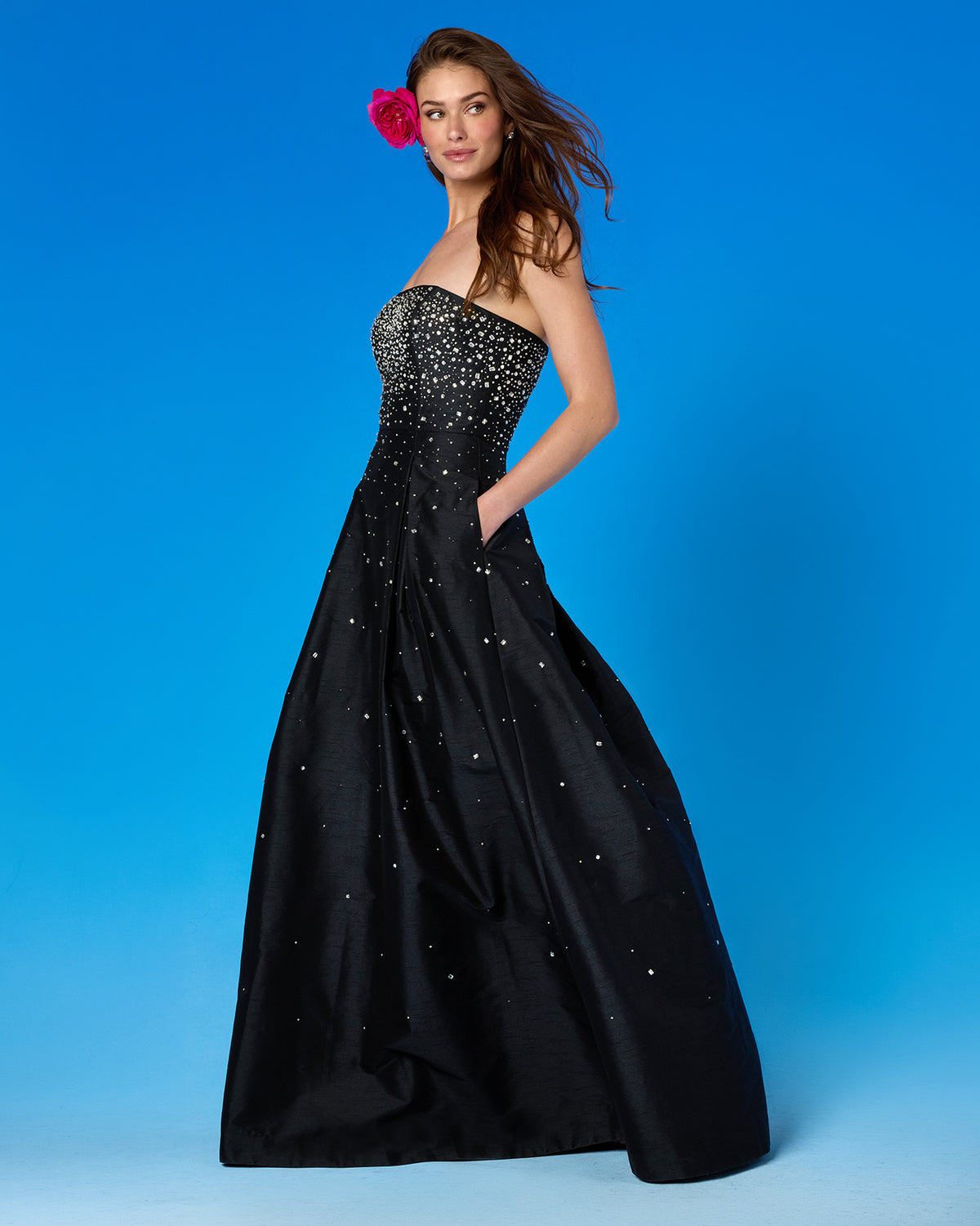 A woman modeling a strapless black formal gown with a heavily embellished beaded bodice and scattered rhinestone-like accents on the full skirt, posed against a bright blue studio background.