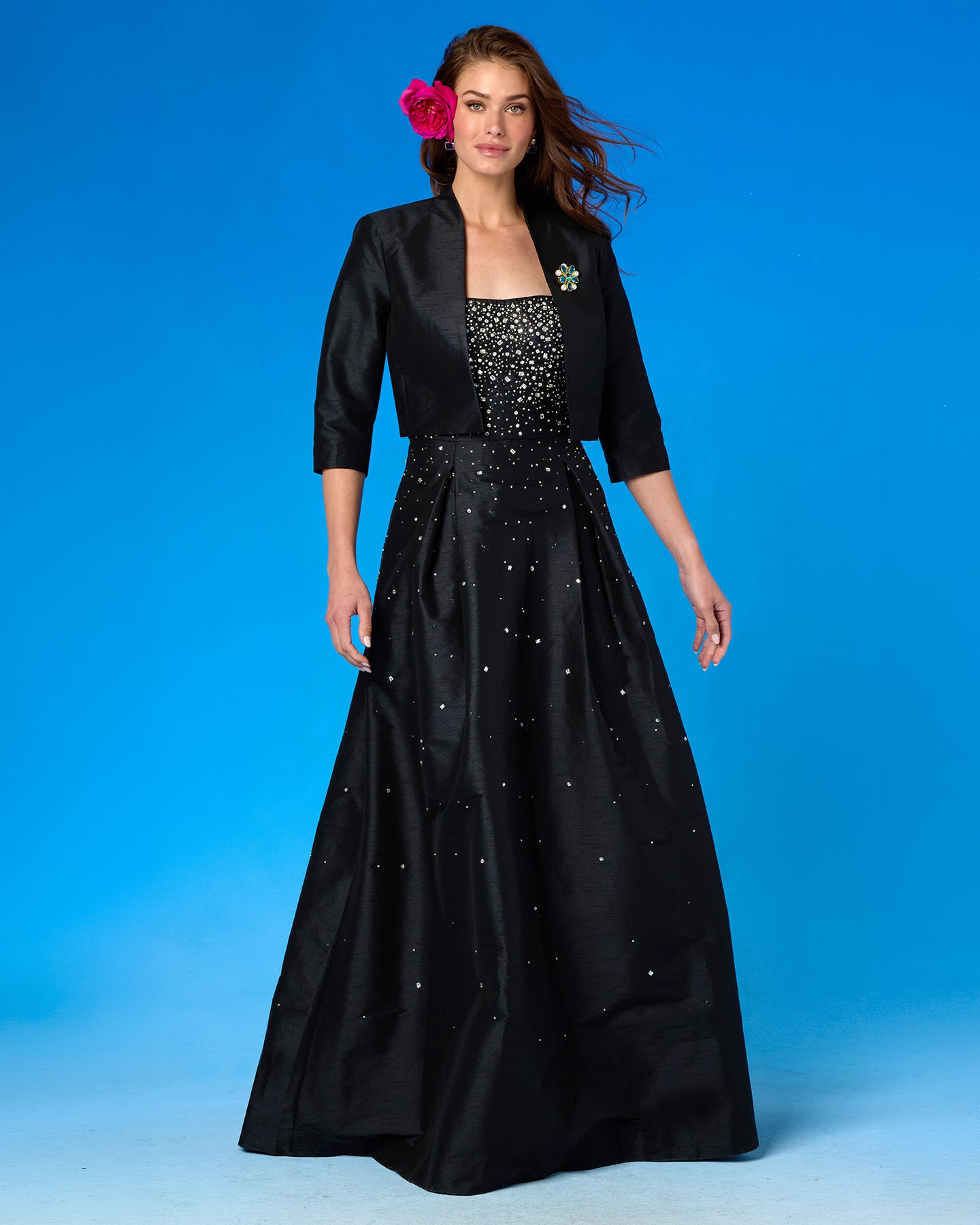 A woman modeling a formal black gown with a matching three-quarter sleeve bolero jacket. The dress features a beaded bodice and scattered rhinestone embellishments on the skirt, set against a solid blue studio background.