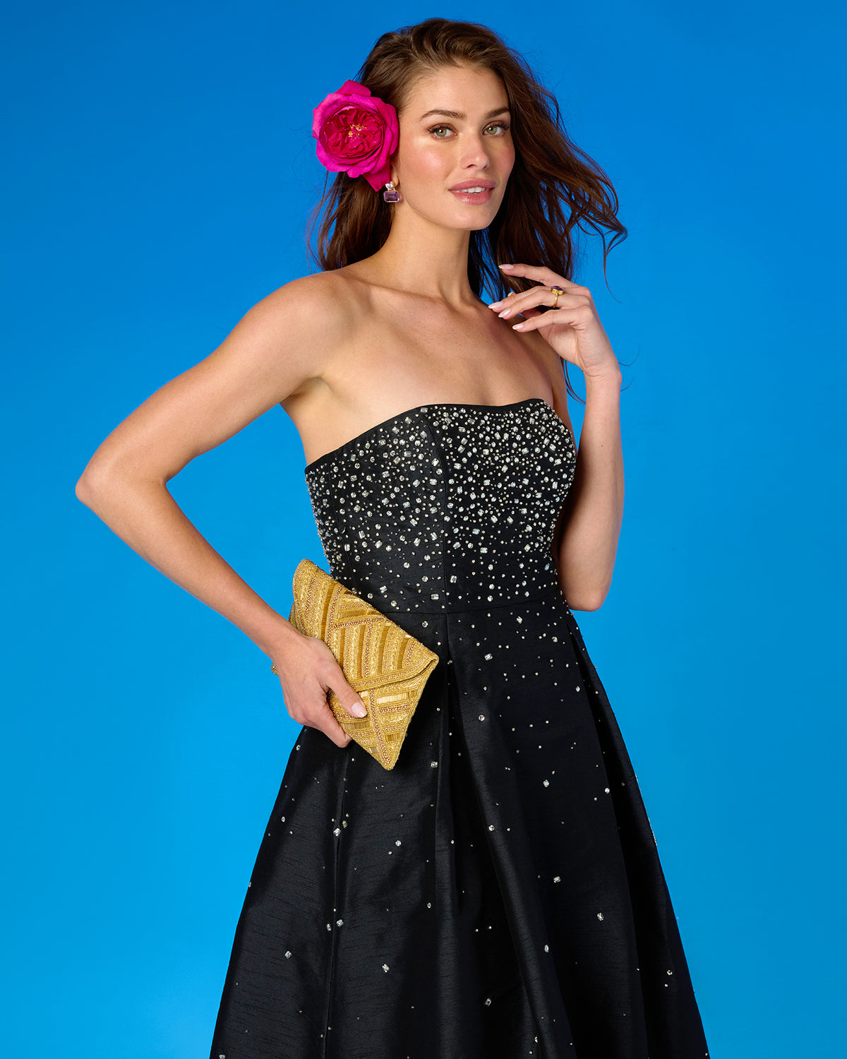 A woman modeling a strapless black tulle gown with silver rhinestone embellishments, holding a gold clutch against a bright blue studio background.
