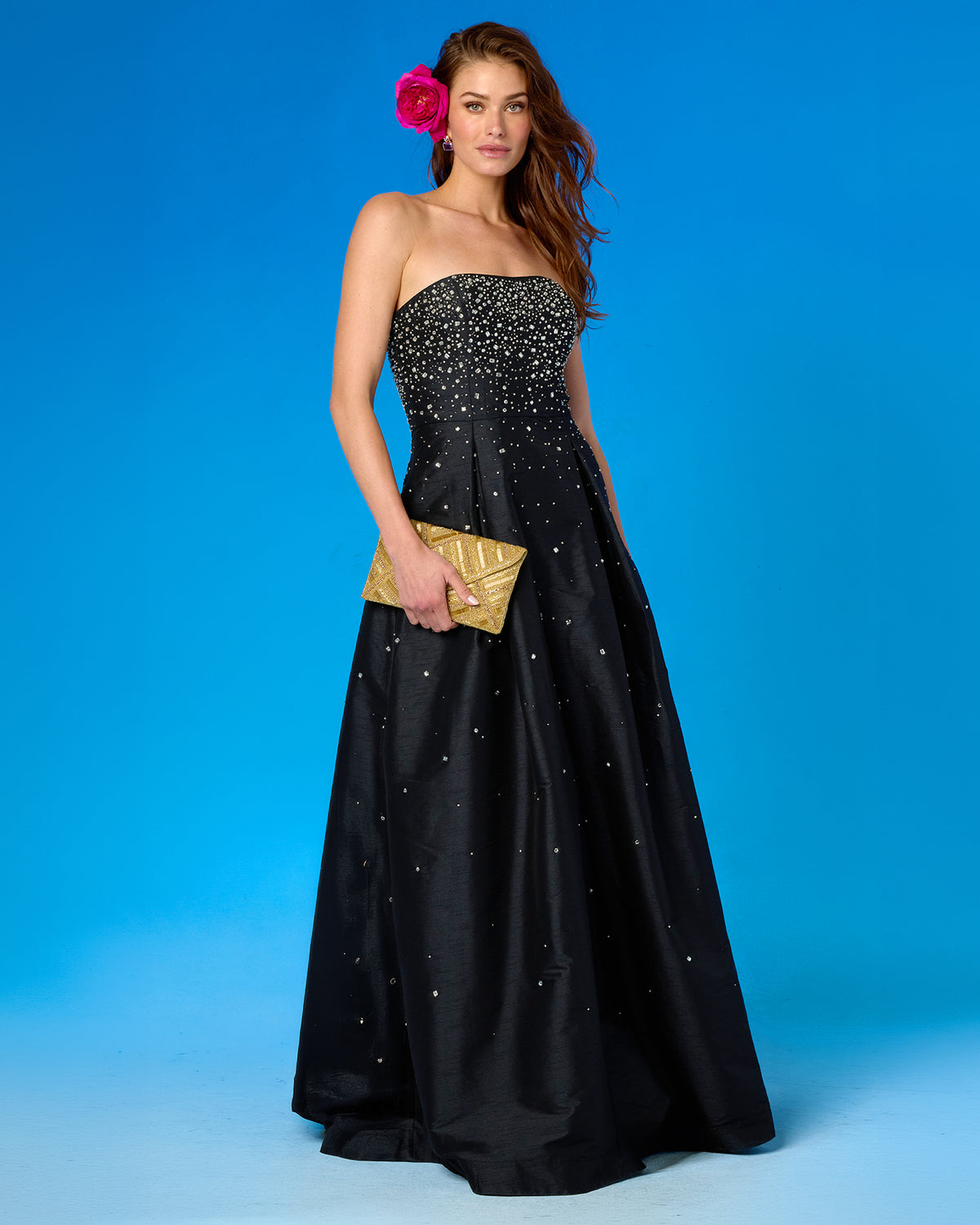 A woman modeling a strapless black ball gown with silver rhinestone embellishments, holding a gold clutch against a bright blue studio background.