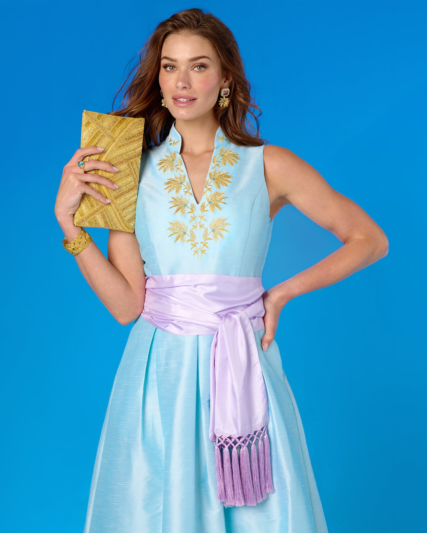 Woman in a light blue dress with gold embroidery and a purple sash against a blue background