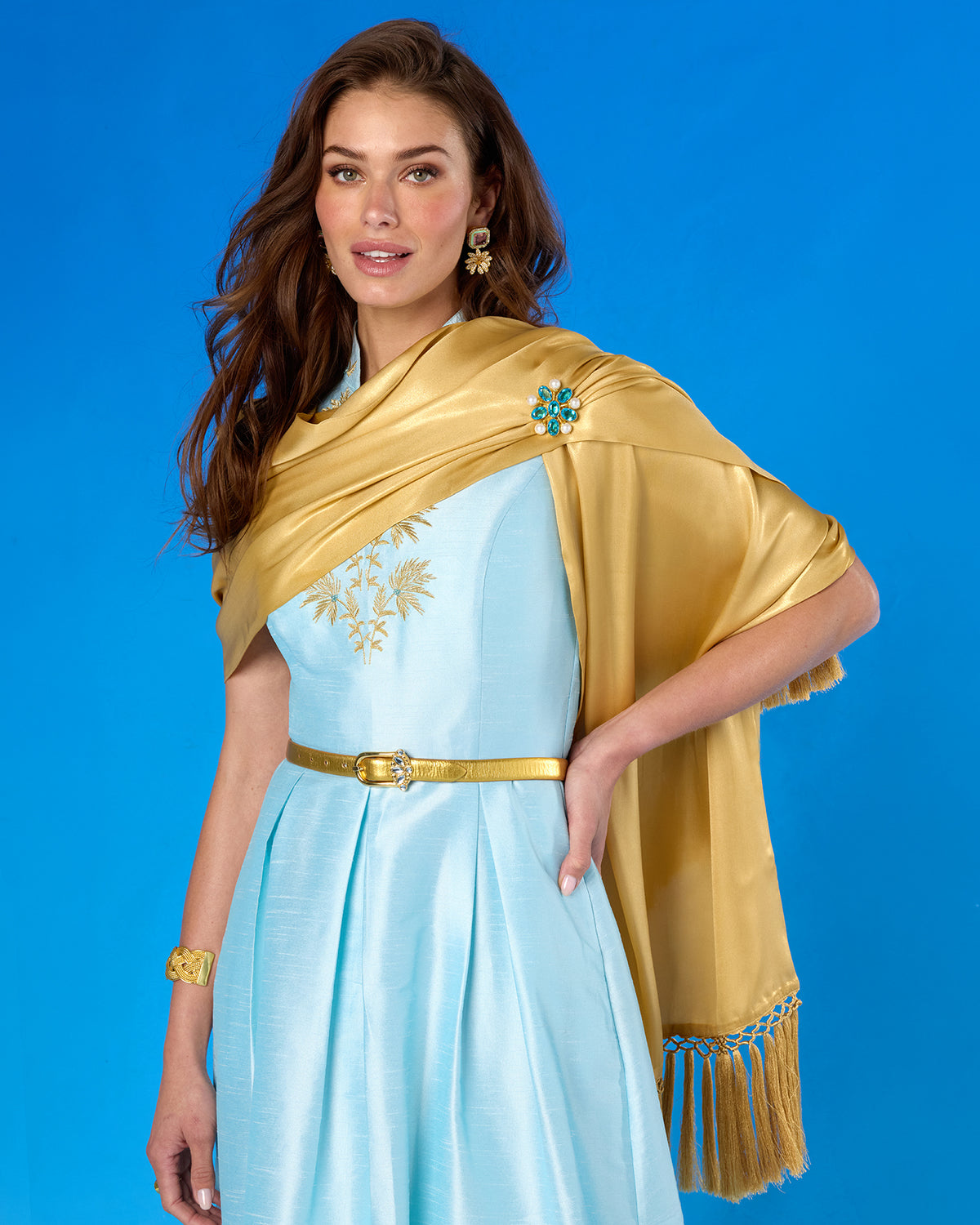 A woman modeling a light blue dress with gold embroidery, a gold belt, and a shiny gold shawl draped over her shoulders, posed against a solid blue studio background.