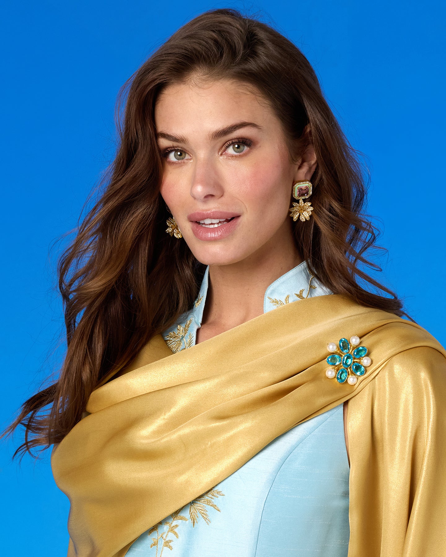 Woman wearing a gold scarf and blue blouse with jewelry against a blue background