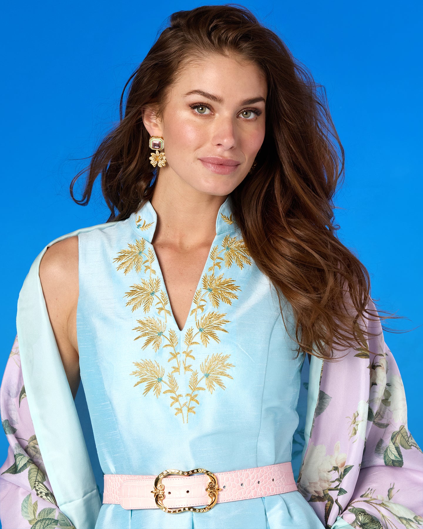 Woman wearing a light blue dress with floral embroidery and a pink belt against a blue background