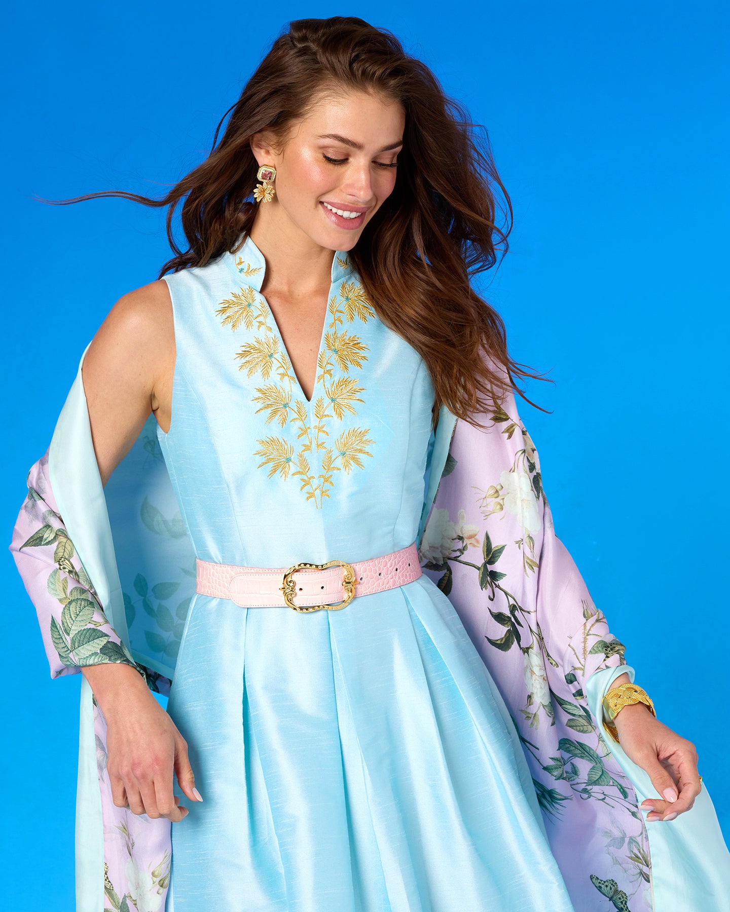 Woman wearing a light blue dress with floral embroidery and a pink belt against a blue background