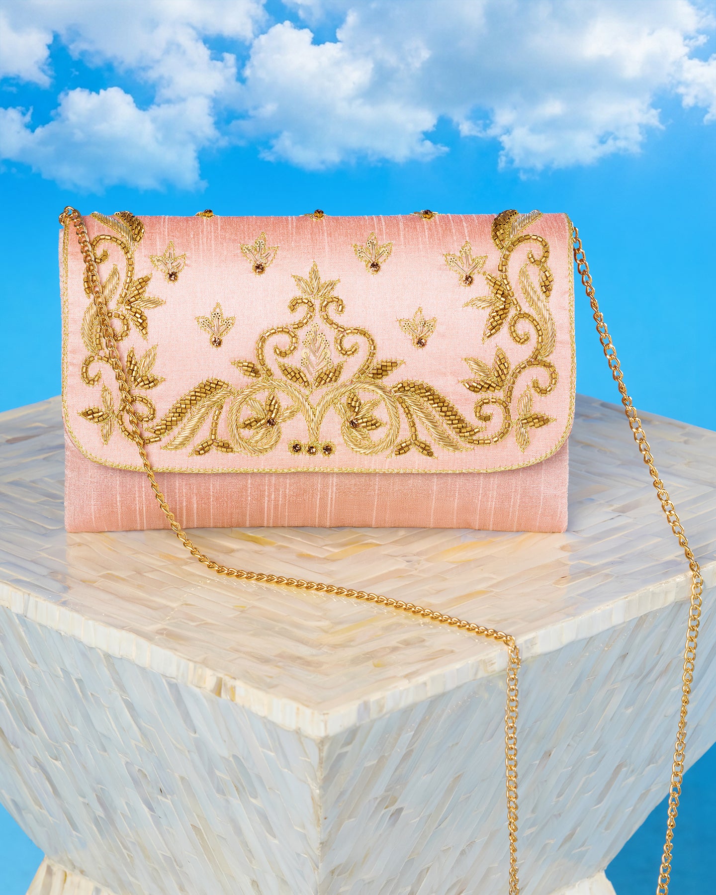 Pink clutch bag with gold embroidery on and chain on a marble surface with a blue sky background