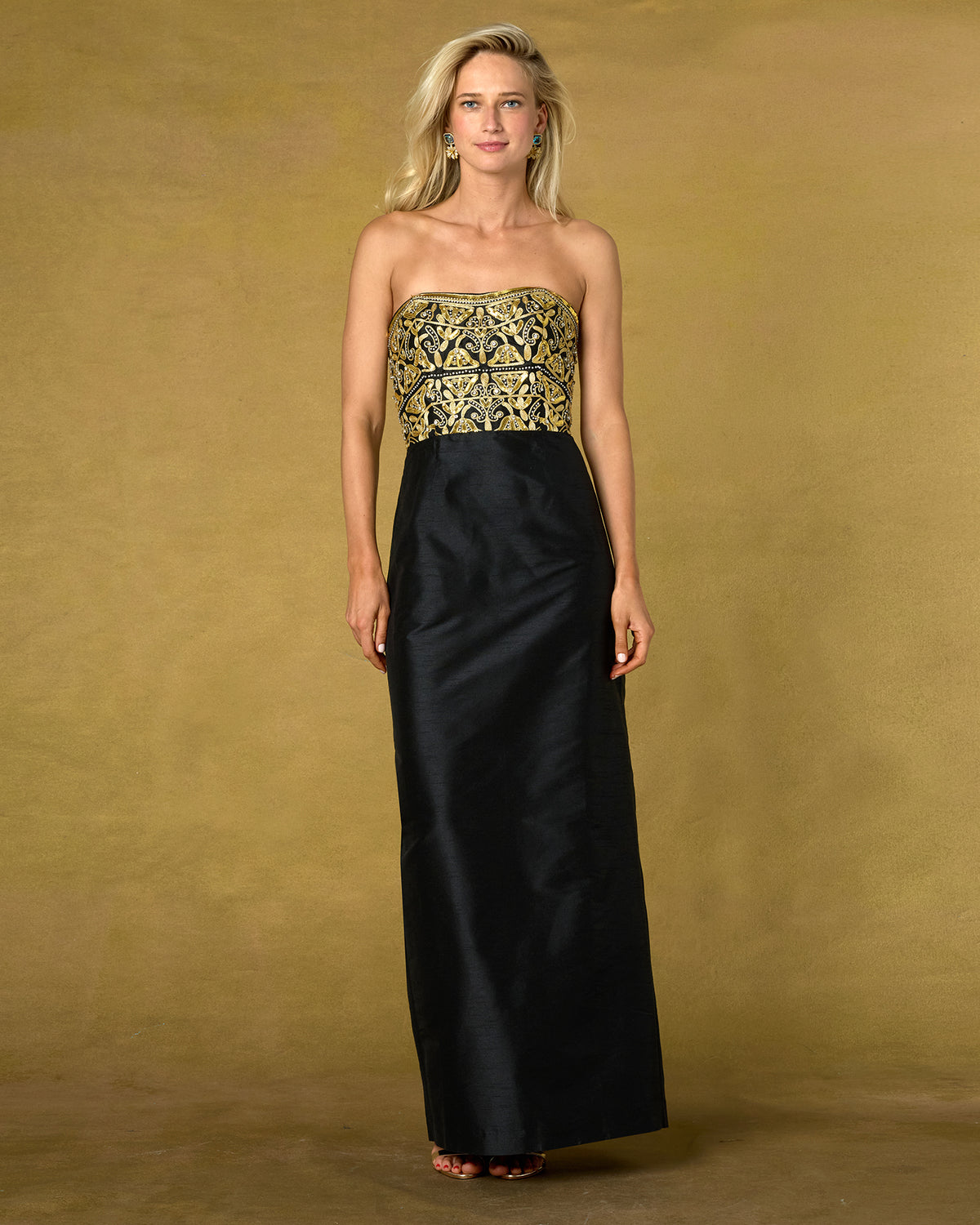 A woman modeling a strapless black maxi gown with an ornate gold embroidered bodice, set against a textured gold studio backdrop.